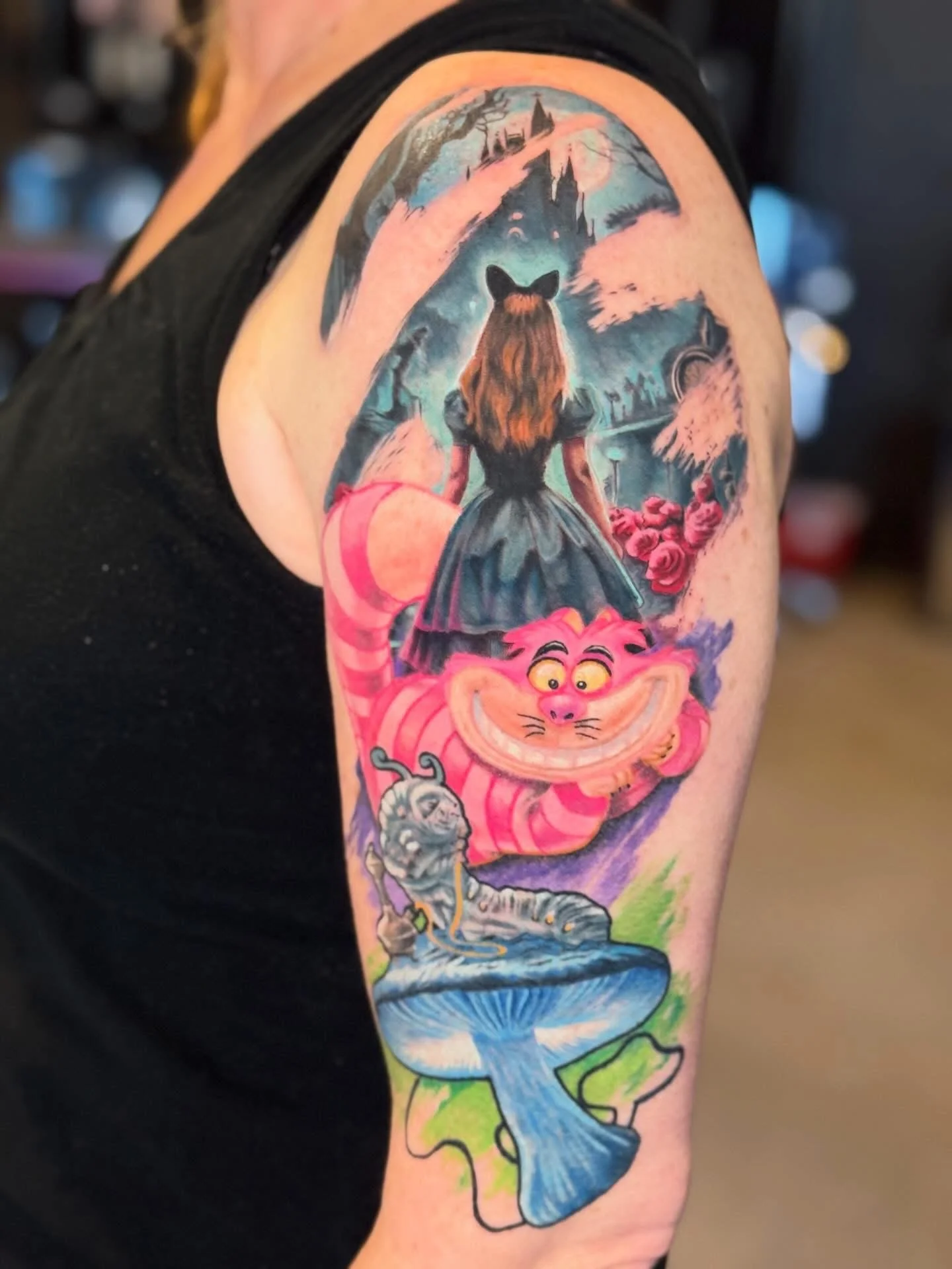 Colorful tattoo of Alice in Wonderland theme on person's upper arm, featuring the Cheshire Cat, a girl with a bear head, pink trees, a castle, and a river with a fish.