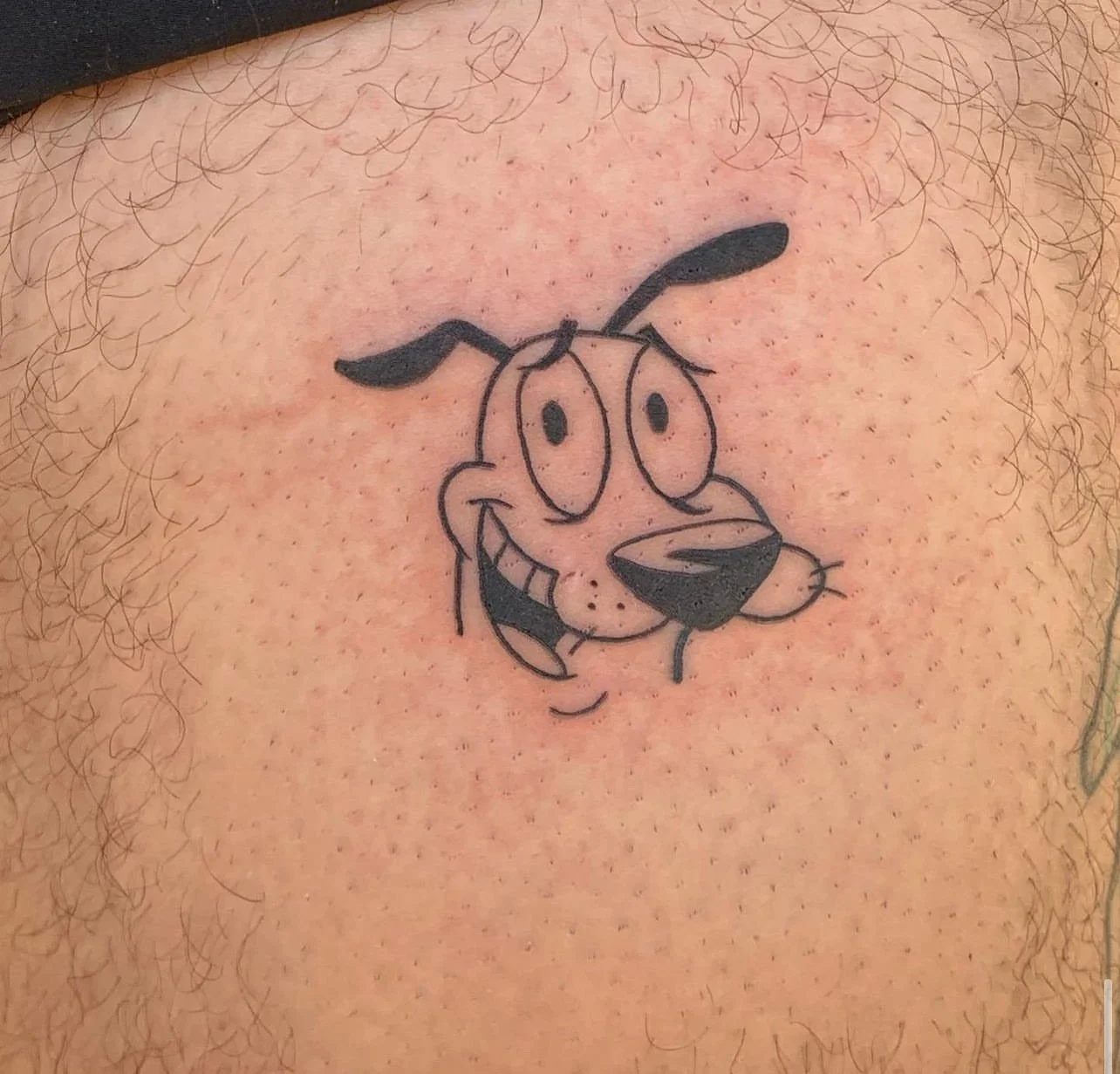 Tattoo of cartoon dog character with floppy ears, big eyes, and a wide smile, located on person's chest with chest hair surrounding it.