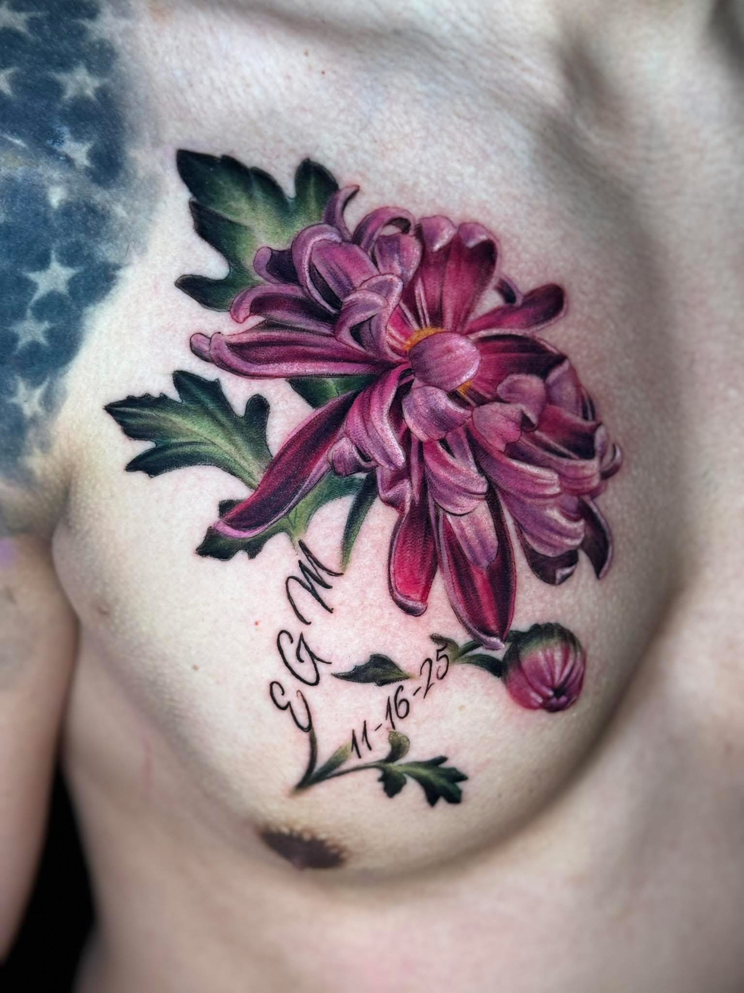 Close-up of a tattoo of a pink chrysanthemum flower with green leaves on person's skin. Text with dates is partially visible beneath the flower.