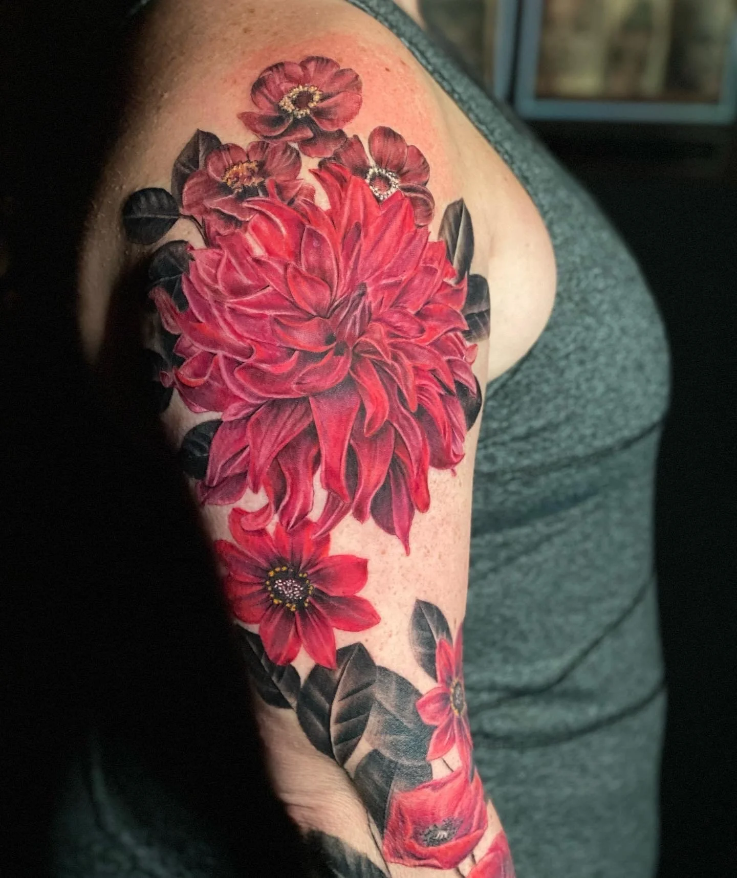 Close-up of a colorful floral tattoo on a person's upper arm, featuring a large red dahlia, smaller red flowers, and dark green leaves.