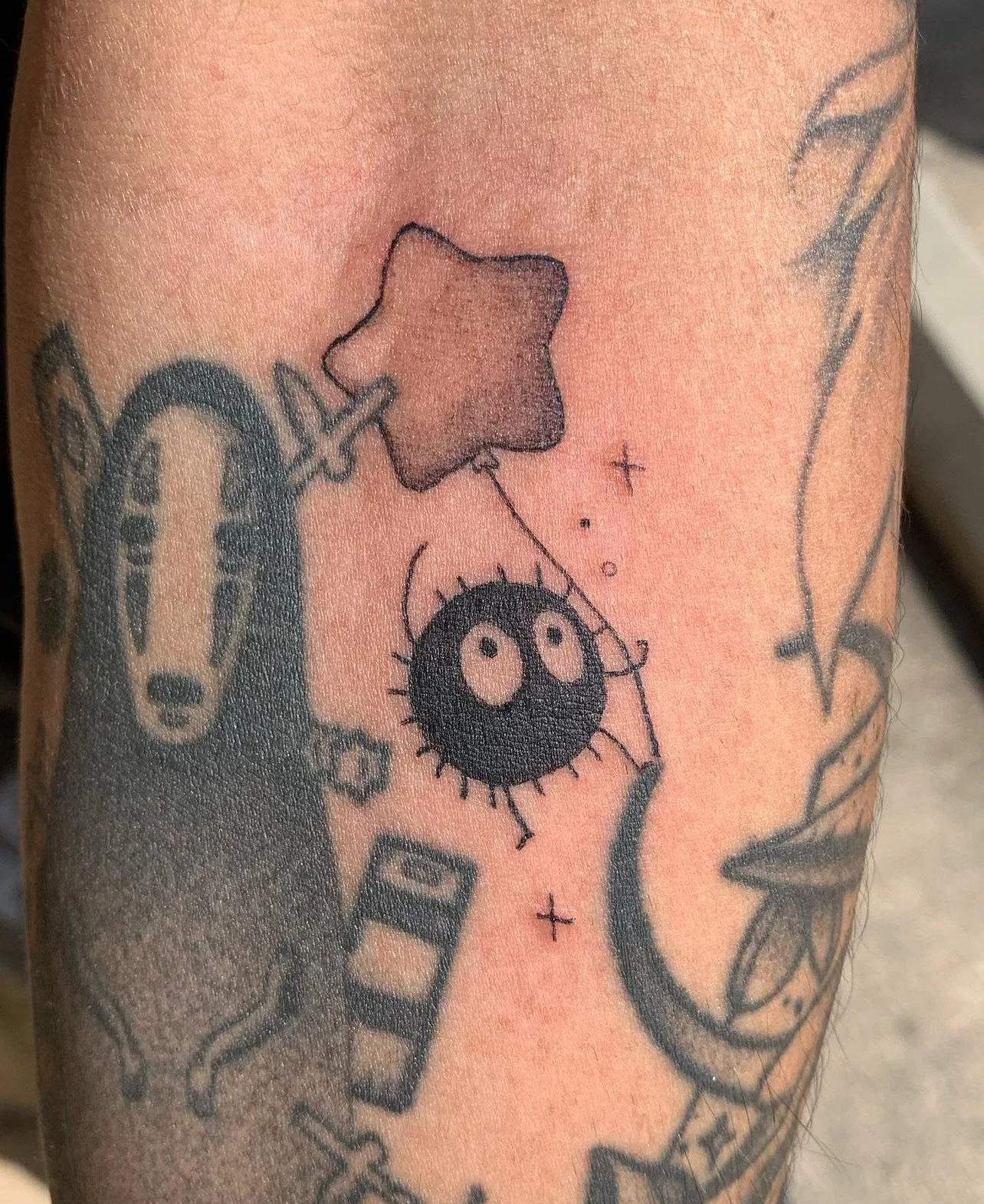 Tattoo of a black fuzzy creature with two big eyes and tiny legs, a star-shaped balloon, a dog face, and a wristwatch on an arm.