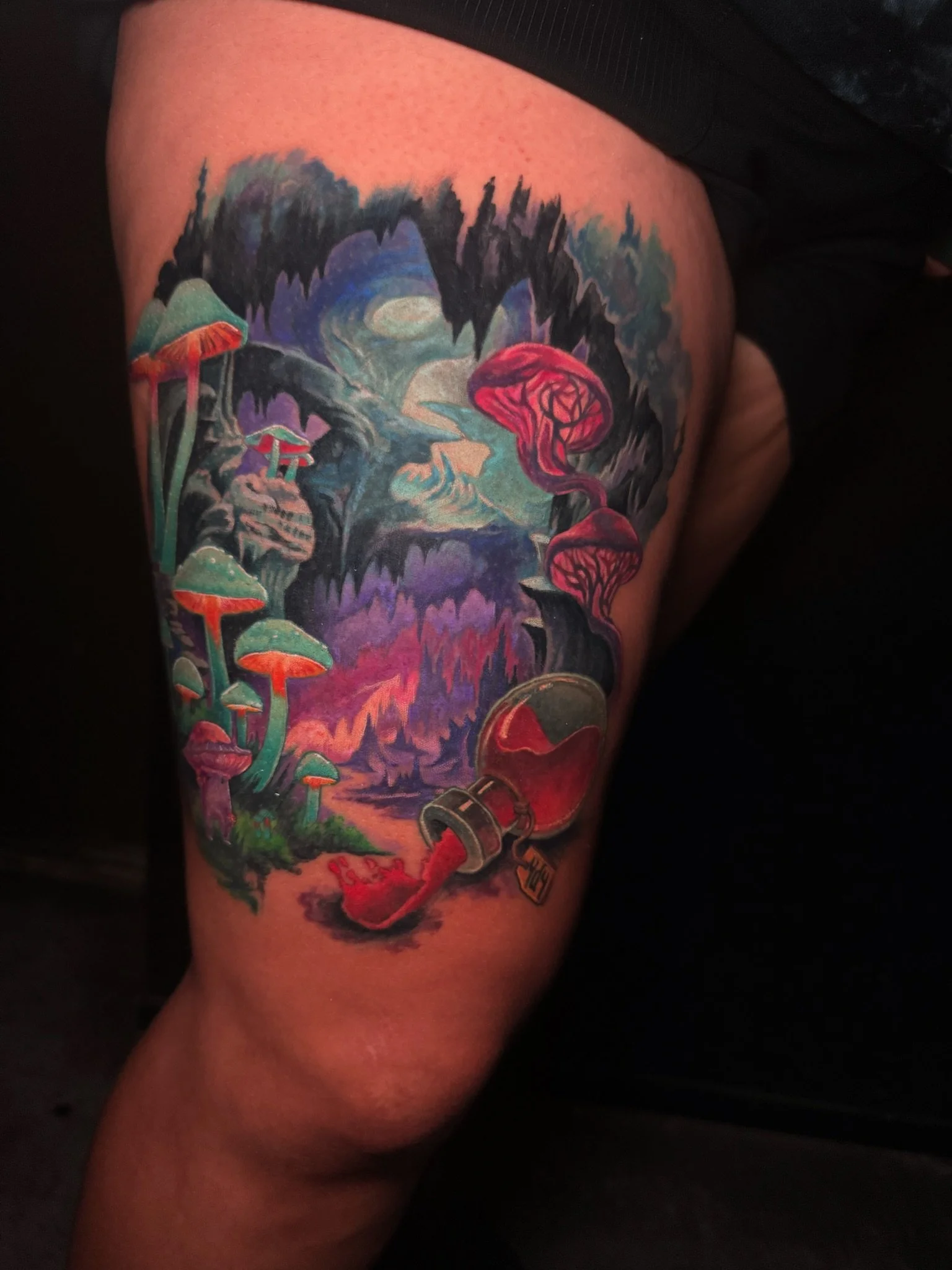 Colorful tattoo on a person's thigh depicting a fantasy scene with mushrooms, a lamp spilling red ink or paint, a cave or forest background, and swirling sky elements.