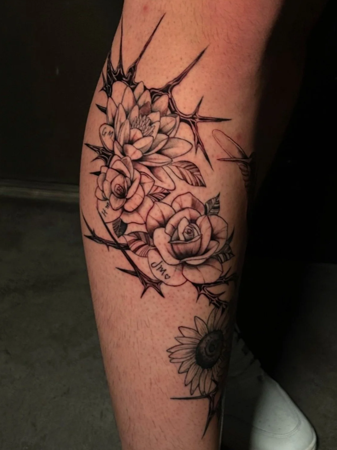 A tattoo on a person's leg featuring detailed black and gray roses, thorns, and a sunflower.