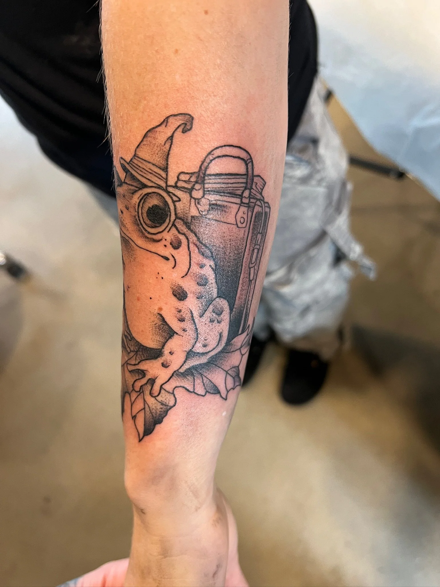 Tattoo of a frog wearing a wizard's hat, with a potion book and bubble effects, on person's forearm.