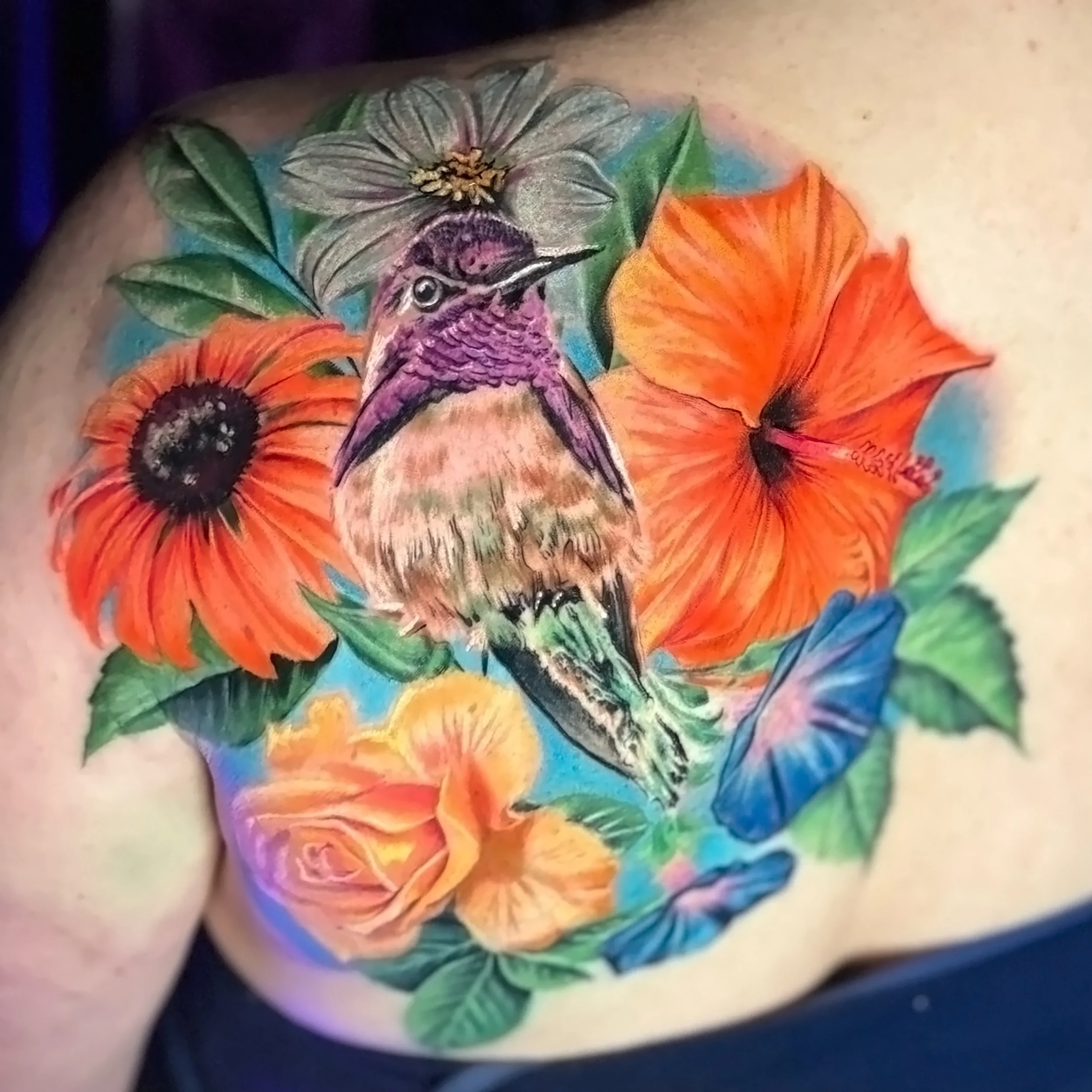 Colorful tattoo of a hummingbird with purple and pink feathers, surrounded by bright orange, yellow, blue, and green flowers with detailed petals and black centers on a person's arm.