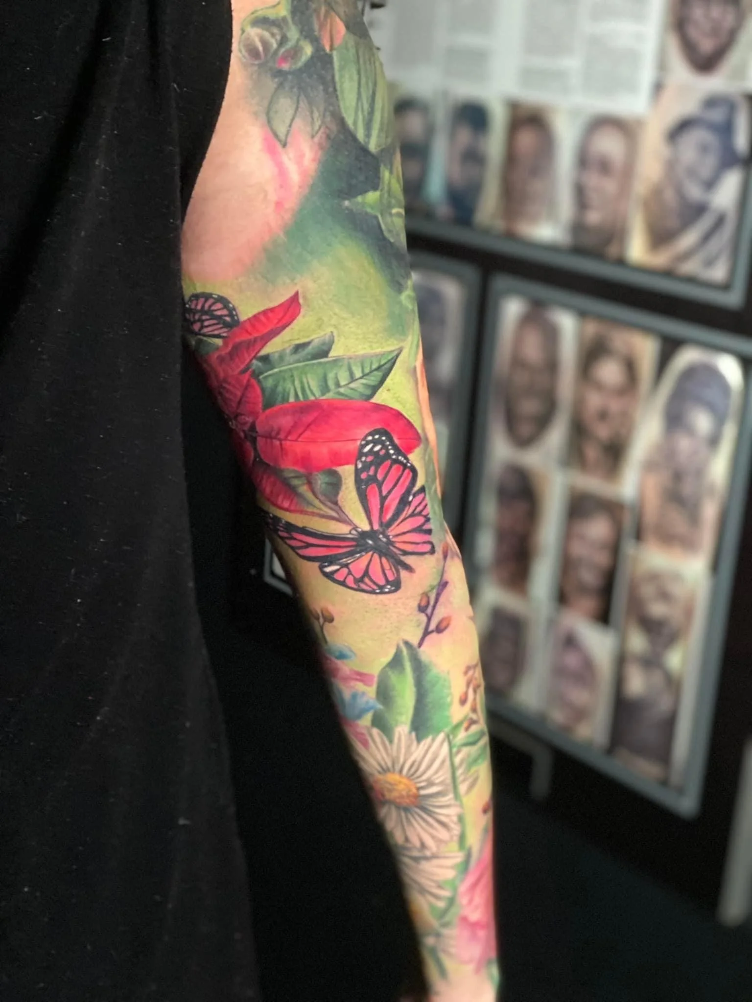 Colorful tattoo sleeve featuring a butterfly, red tropical leaves, green foliage, and a white daisy on a person's arm with a black sleeve.