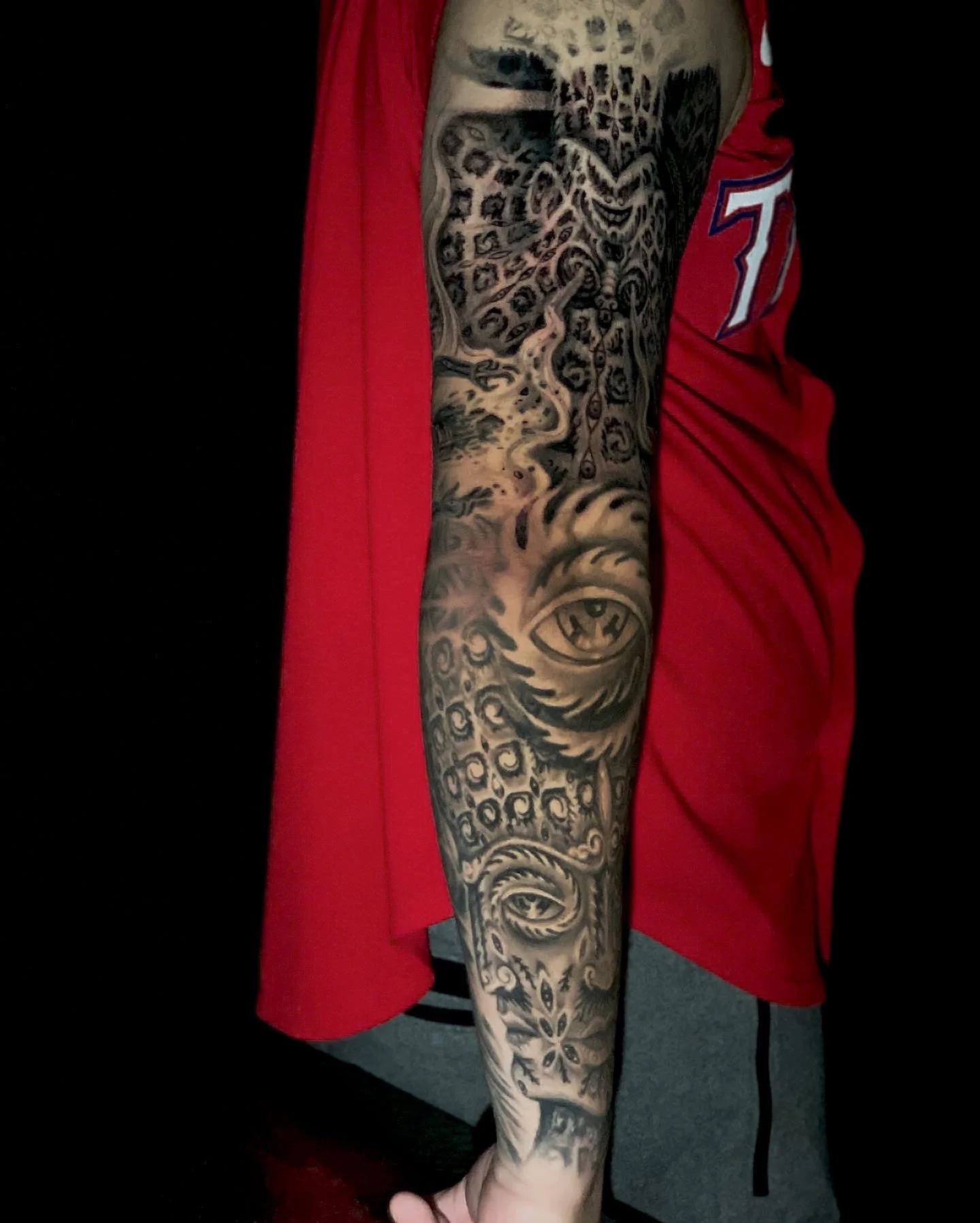 A person's arm with a detailed black and gray tattoo sleeve featuring an animal face, swirling patterns, and abstract designs, wearing a red sports jersey.