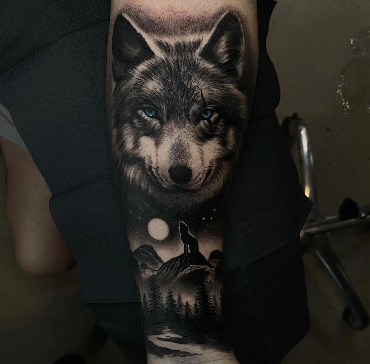 Tattoo of a realistic wolf's face with blue eyes, howling at a full moon in a forested landscape under a starry sky.