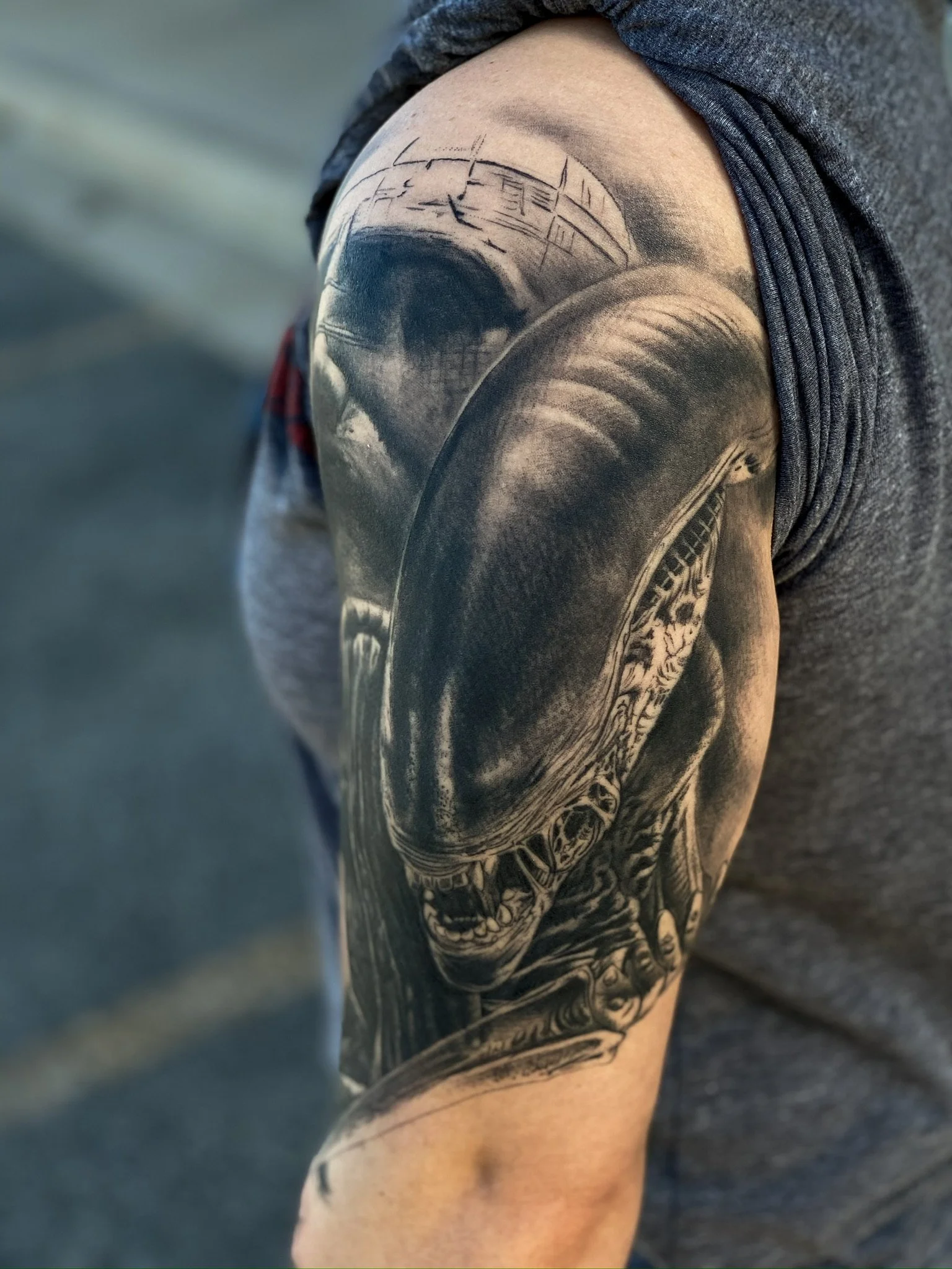 Black and gray tattoo of a detailed alien creature or predator face on a person's upper arm and shoulder.