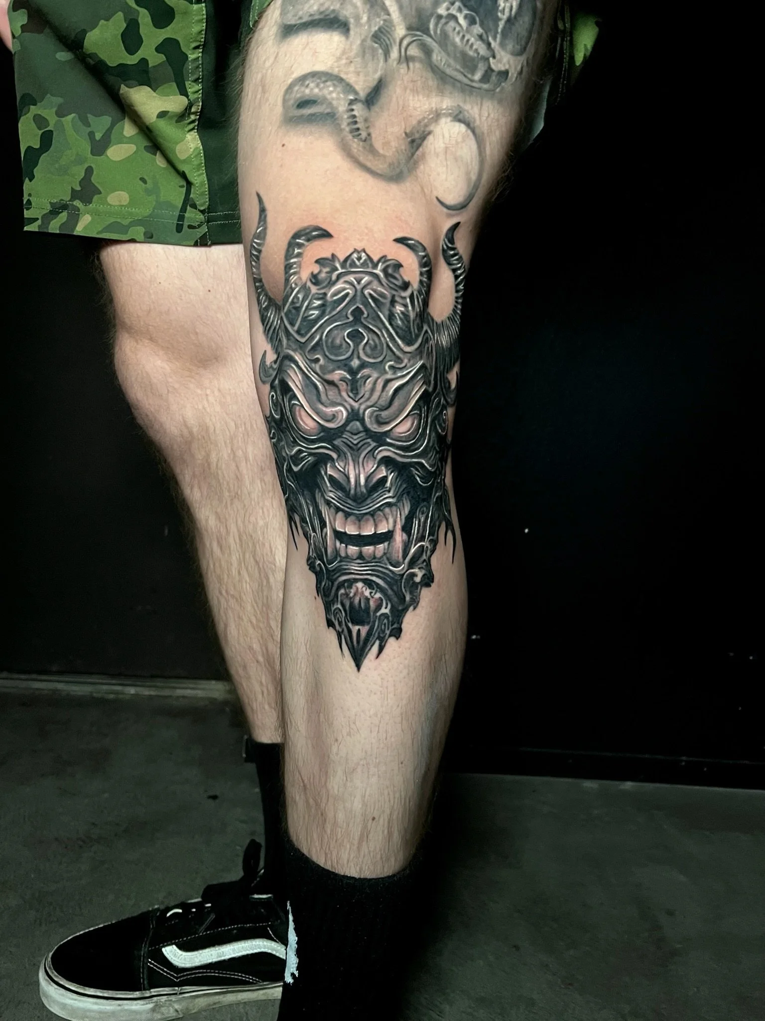 Tattoo of a menacing, stylized demon face with horns on the person's upper leg, in black and gray ink.
