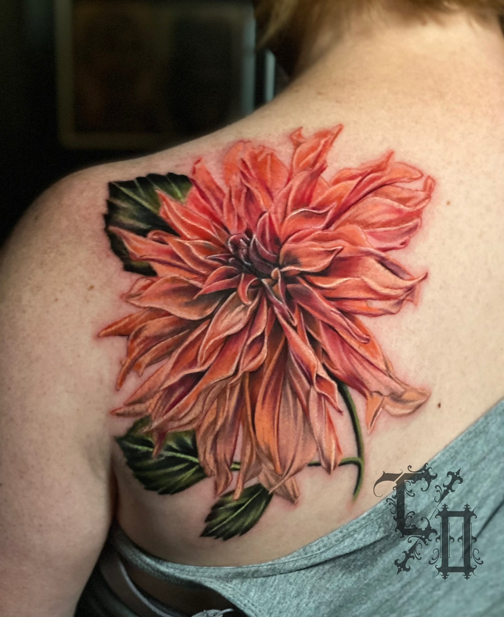 Colorful tattoo of a large orange dahlia flower with green leaves on a person's shoulder.