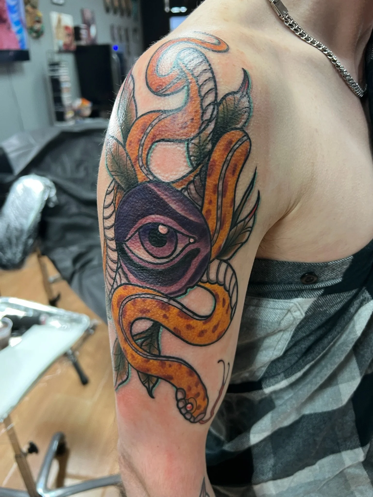Tattoo of a purple eye and a yellow-orange snake with black spots, intertwined with leaves and swirling lines on a person's upper arm and shoulder.