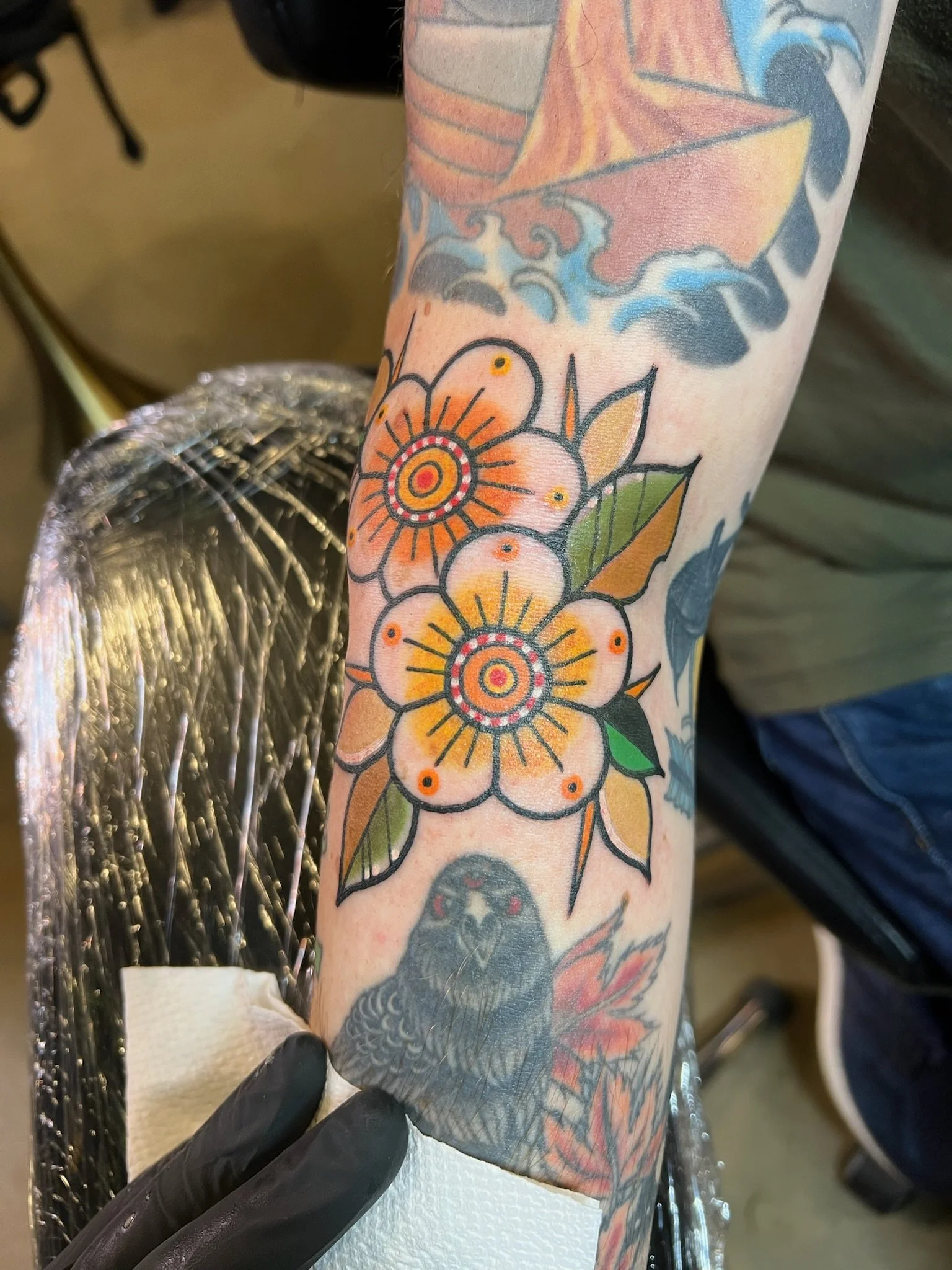 Tattoo of orange and yellow flowers with green leaves on a person's arm, featuring detailed black outlines and dotted center patterns, with additional tattoos visible in the background.