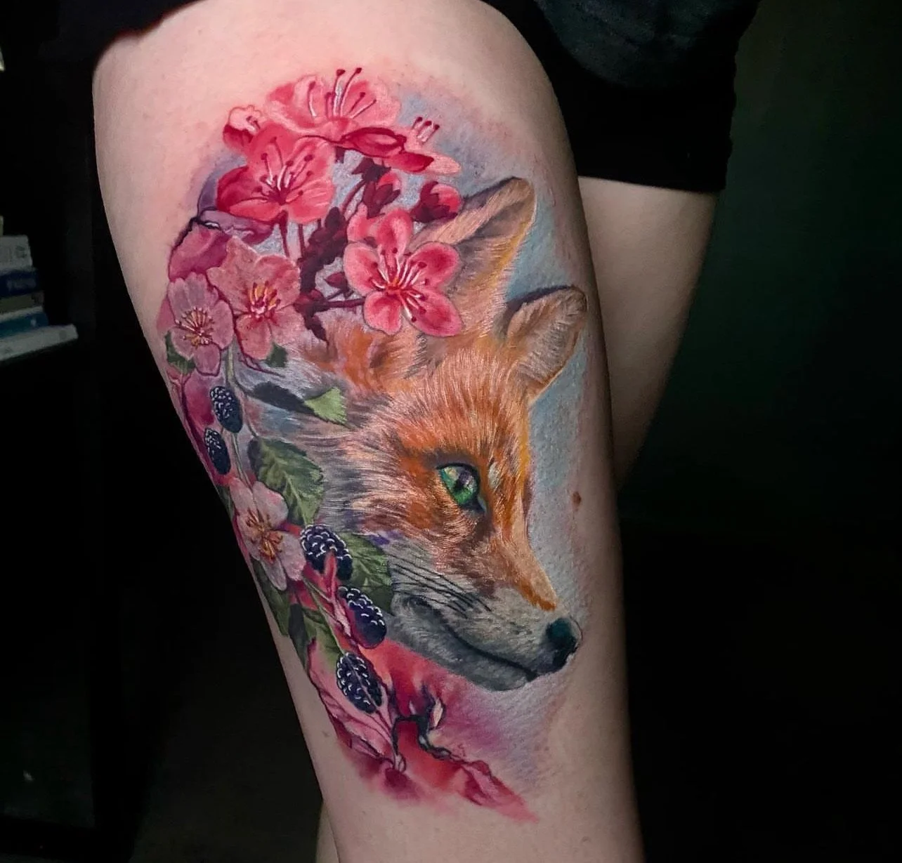 Colorful tattoo of a fox surrounded by pink flowers and blackberries on a person's thigh.