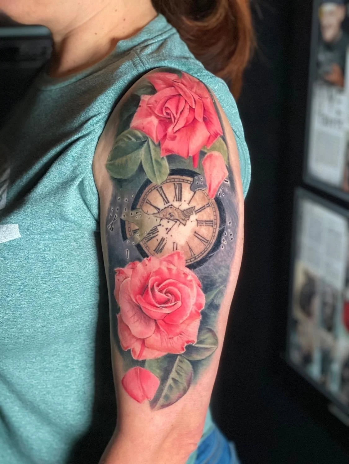 A tattoo on a person's upper arm features two pink roses, green leaves, a vintage clock with roman numerals, and hummingbirds near the clock.