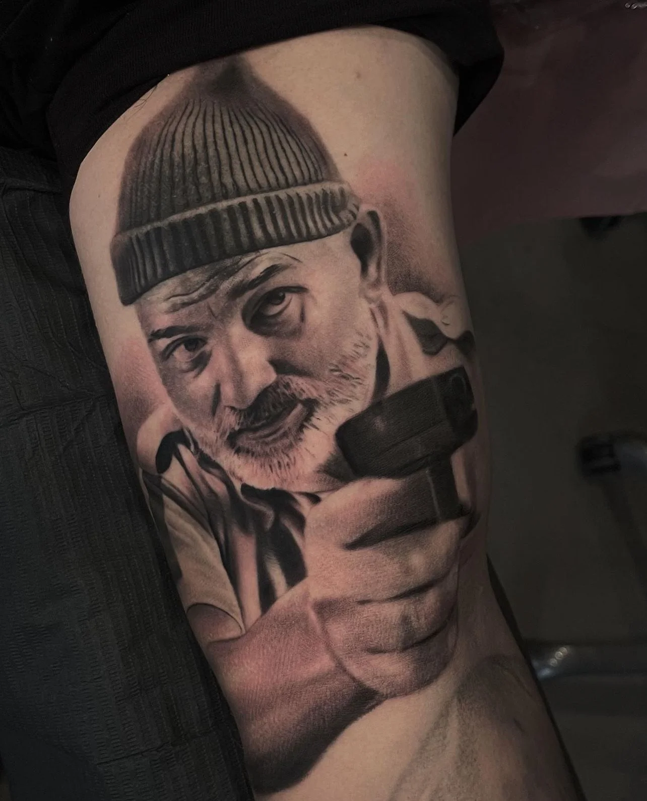 Black and gray tattoo of an elderly man wearing a beanie hat, holding a camera.