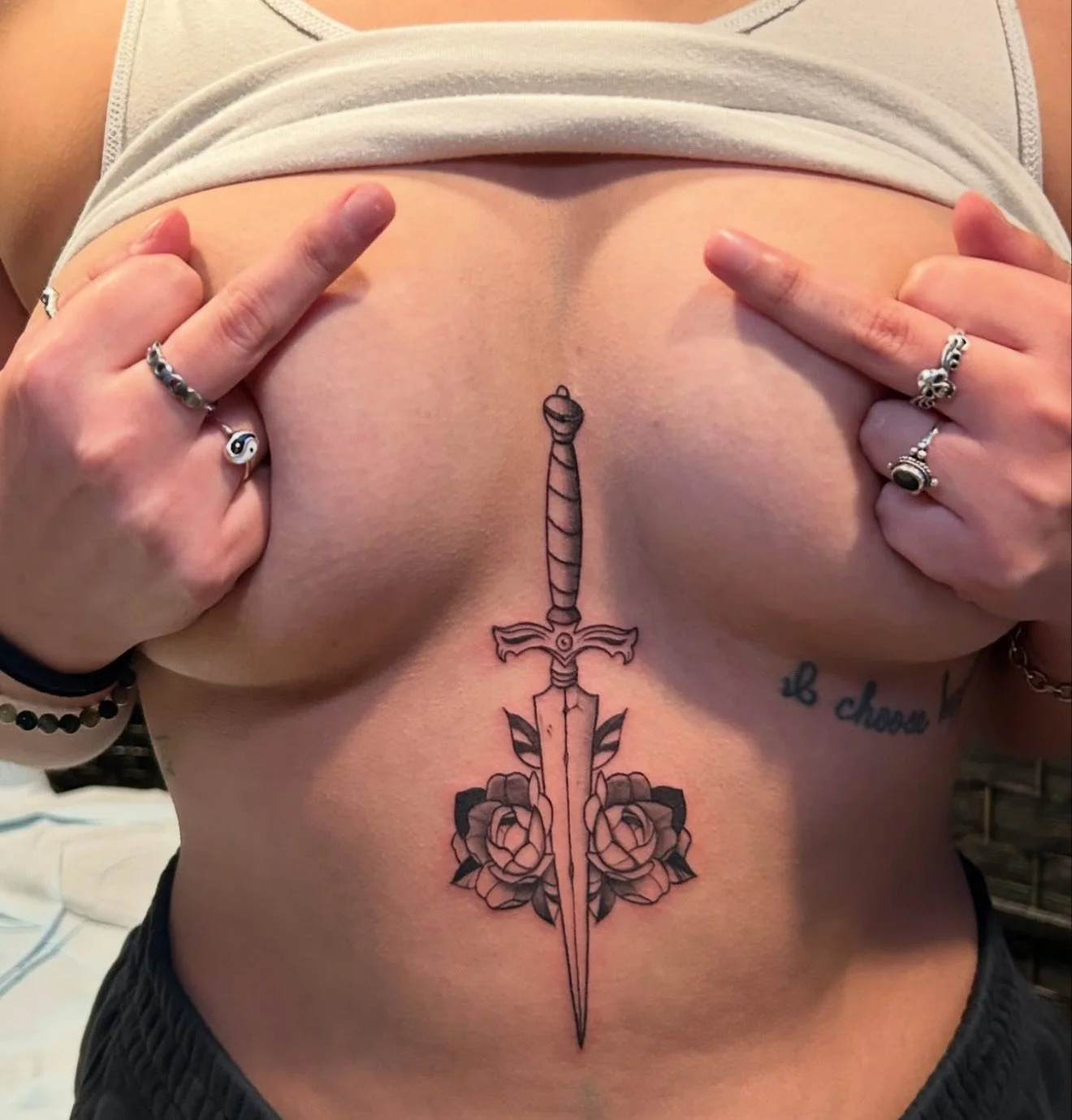 Tattoo of a sword with roses on the person's abdomen, with hands pointing to the tattoo.