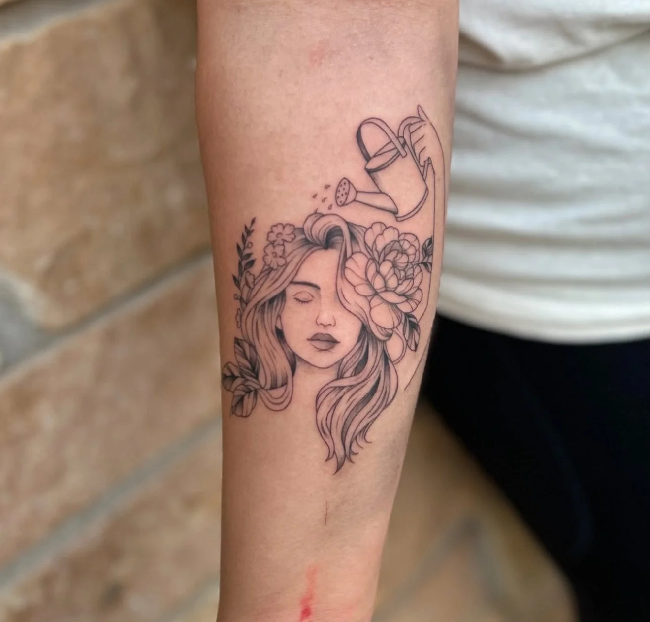 Tattoo of a woman with long flowing hair and flowers in her hair on her forearm, along with a line drawing of a watering can pouring water above her head.
