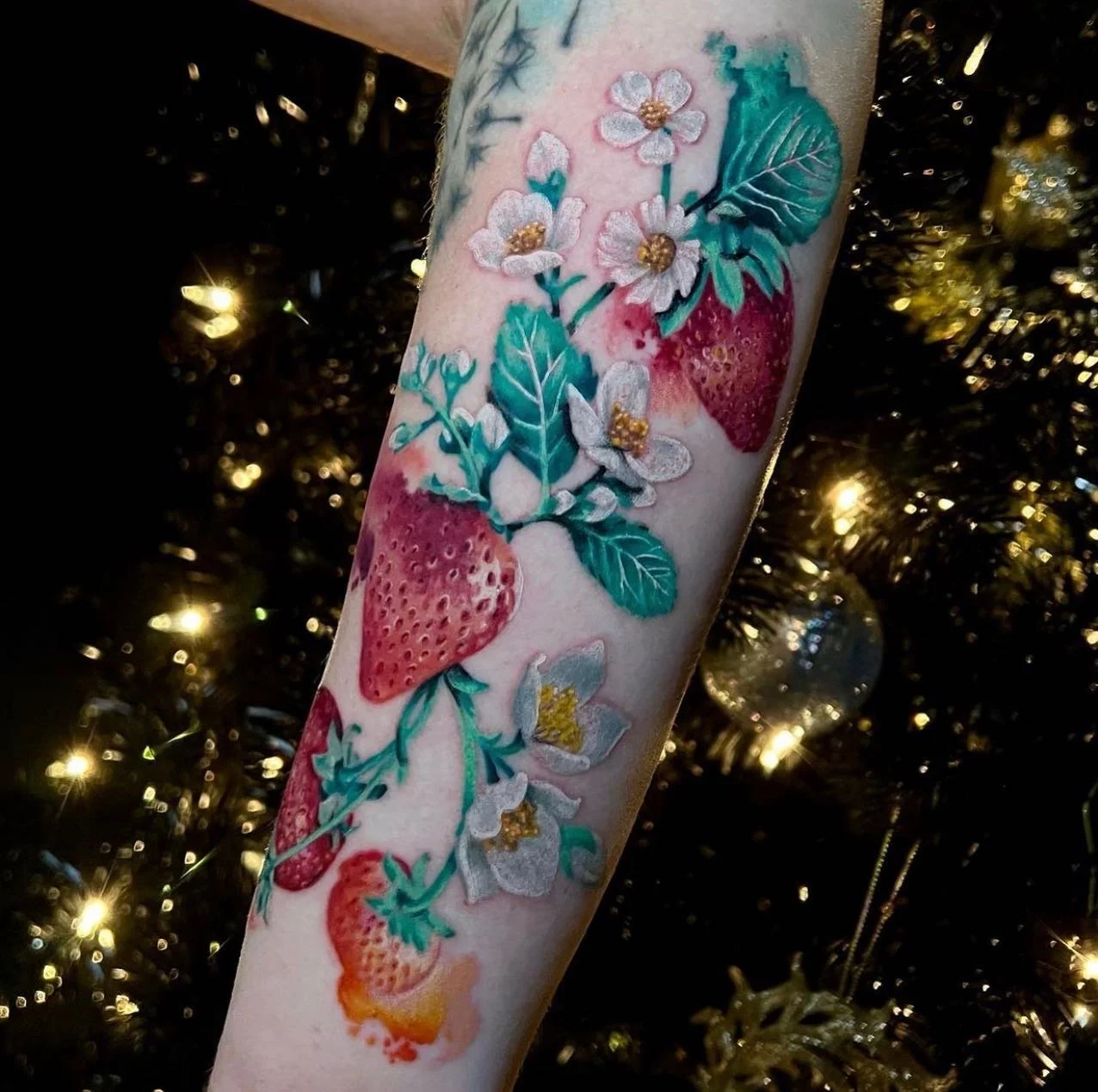 Tattoo of strawberries, white flowers with yellow centers, dark green leaves, and teal foliage on forearm, with a Christmas tree with gold lights in the background.