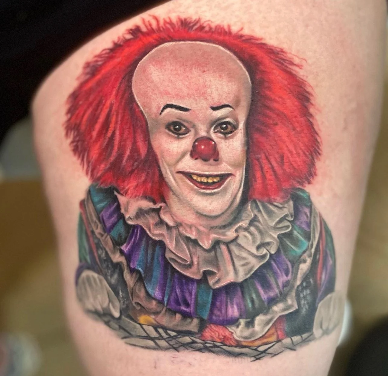 Tattoo of a clown with red hair, bald head, clown nose, and makeup. Wearing a colorful ruffled collar and clown costume.