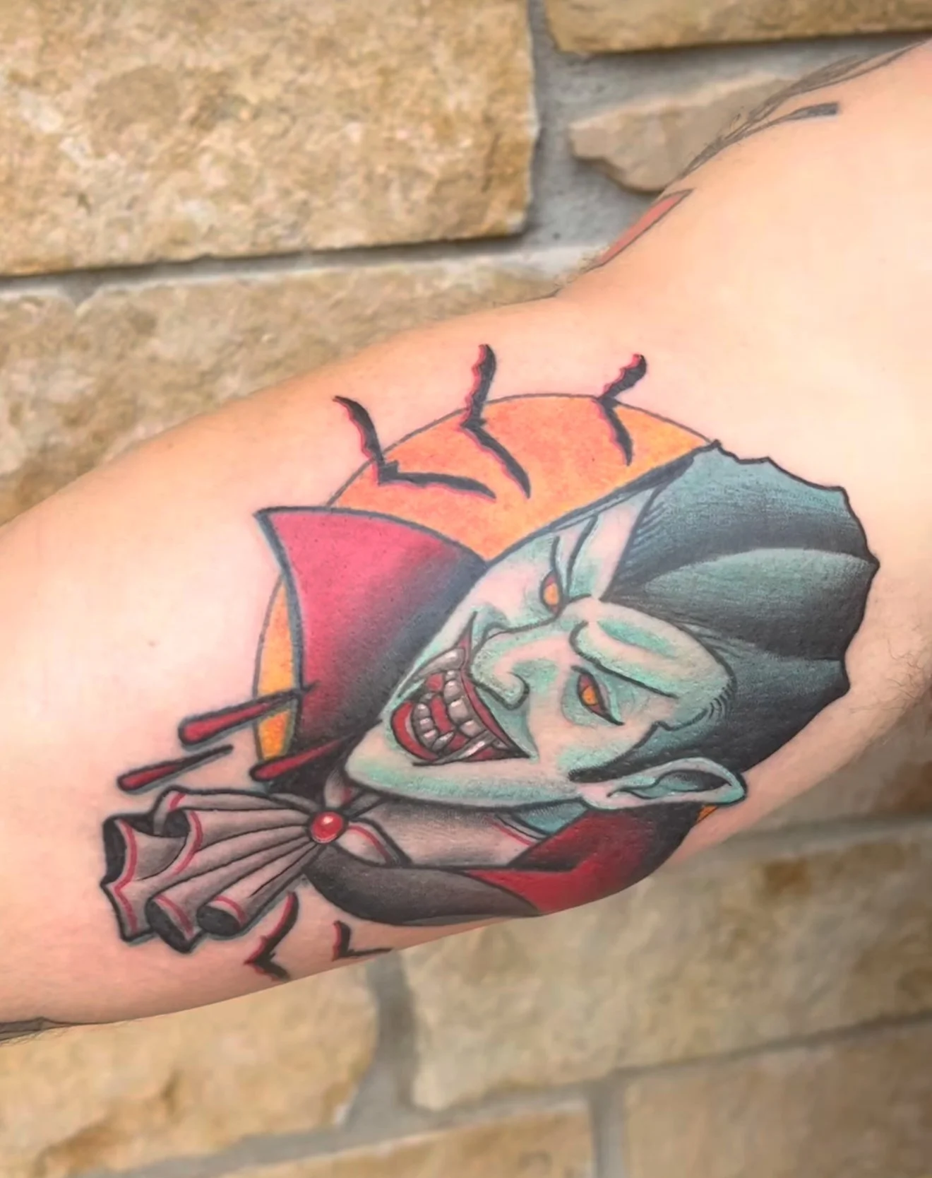a colorful tattoo of a vampire with a yellow moon behind him and a traditional red and black cape with blood dripping, bats in the background on someone's inner bicep