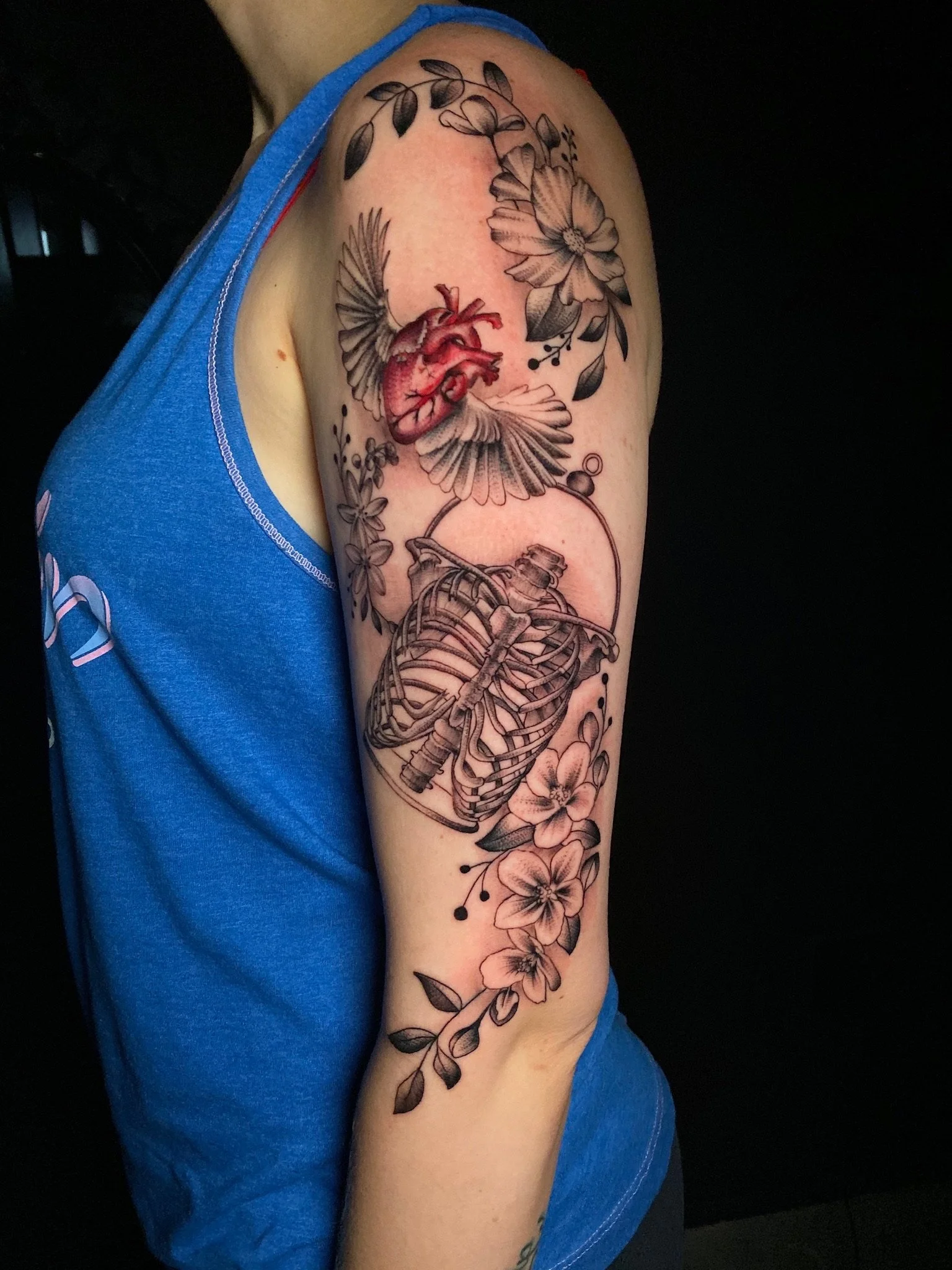 Tattoo of a human heart, a ribcage, and flowers with leaves on a person's upper arm.