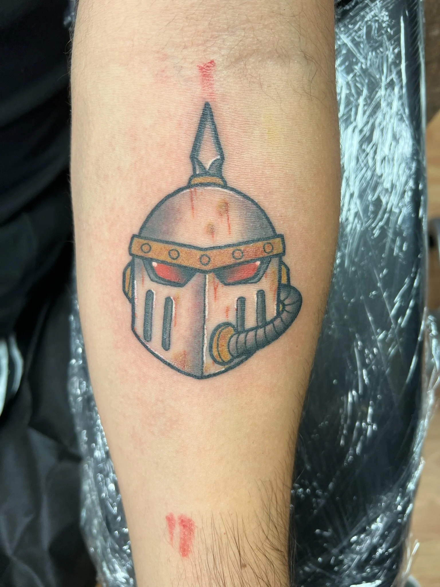 Tattoo of a medieval helmet with red eye slits and a breathing tube on a person's arm.