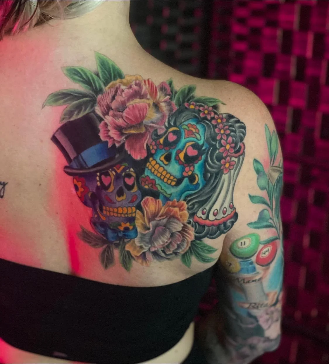 Colorful tattoo of a sugar skull, woman with flowers, and traditional Day of the Dead motifs on a person's upper chest and arm.