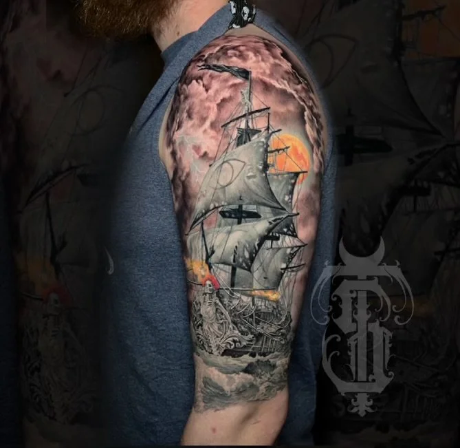 Tattoo of a sailing ship at sea with a sunset and cloudy sky on a man's upper arm.