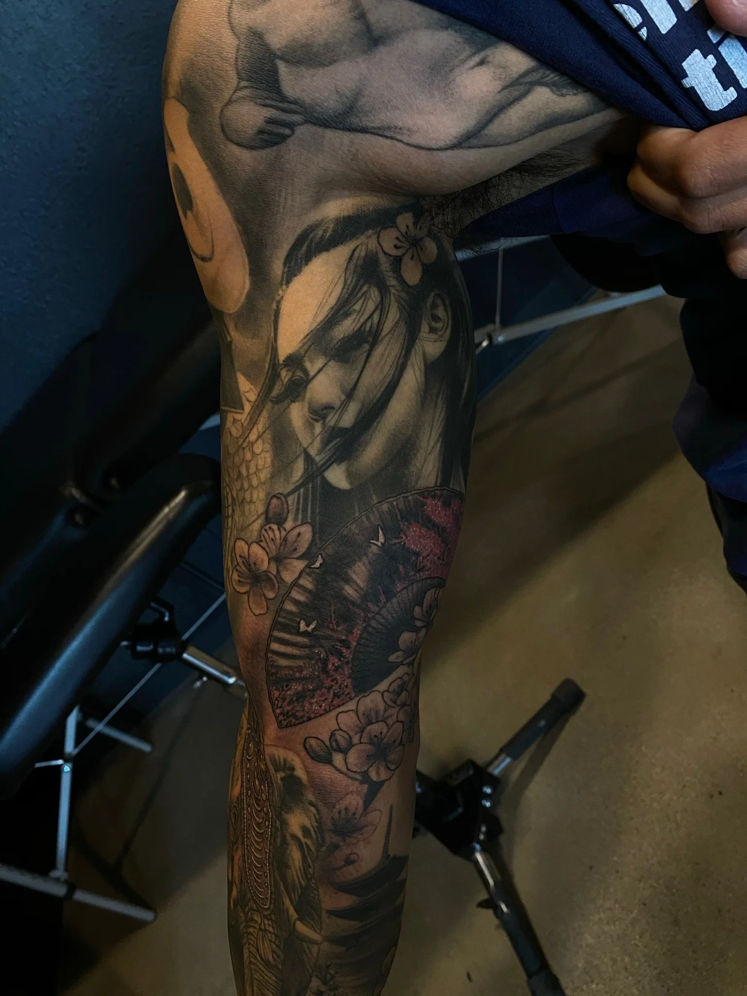 Close-up of a tattooed arm featuring a detailed black-and-gray portrait of a woman with long hair, a flower in her hair, and an ornate fan with floral and butterfly designs surrounding her.