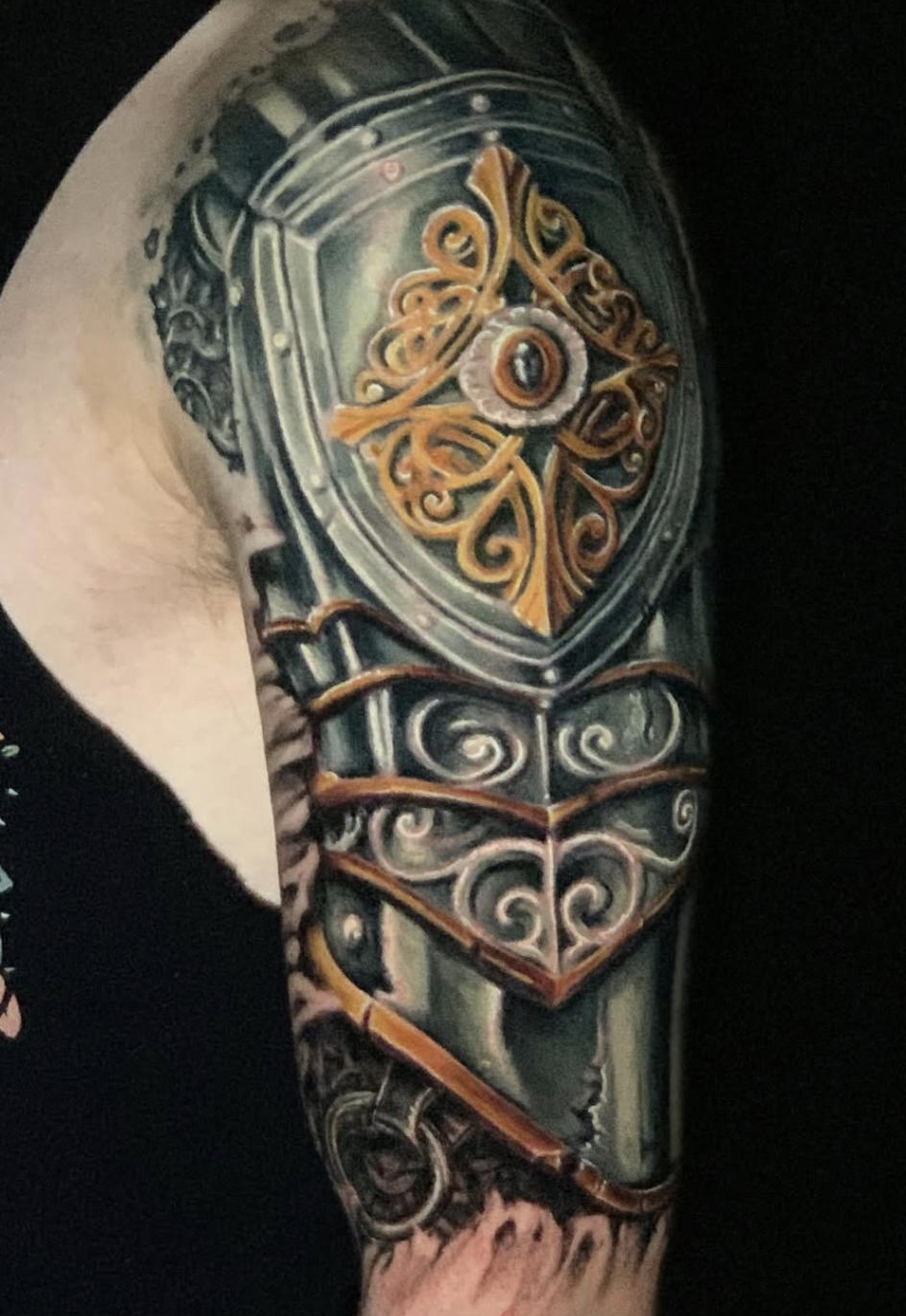 A detailed tattoo on a person's upper arm featuring a sword, a shield with intricate designs, and gold accents with a central eye-like symbol surrounded by ornate scrollwork.