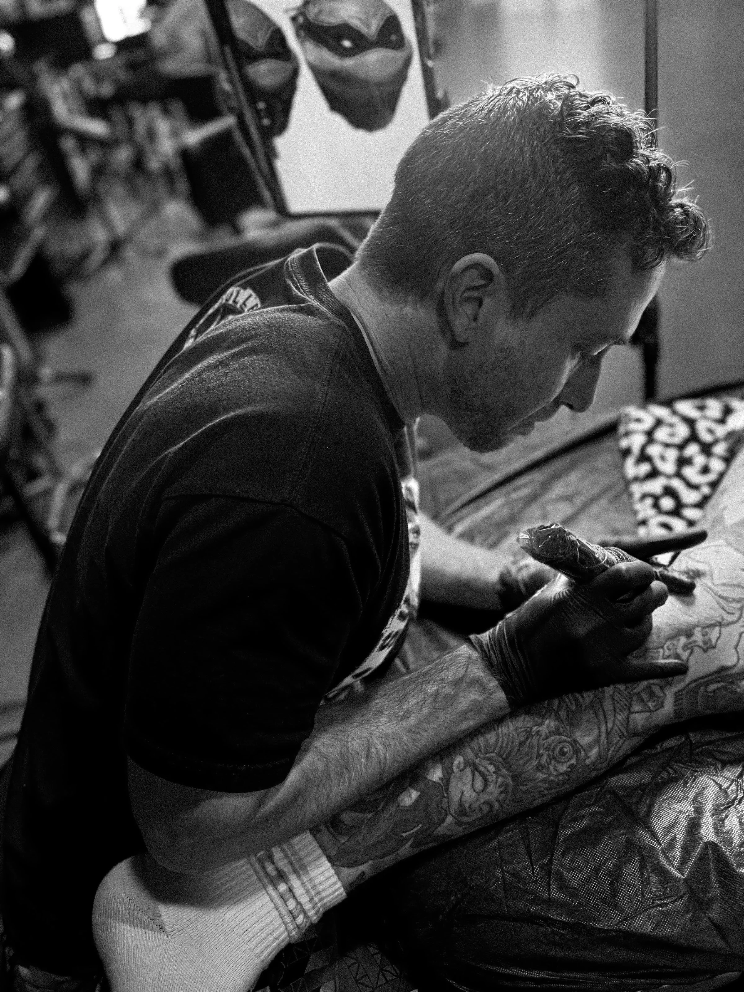 A man with short curly hair and tattoos on his arm is sitting on a chair, leaning forward, and working on a tattoo on another person's leg, using a tattoo machine.