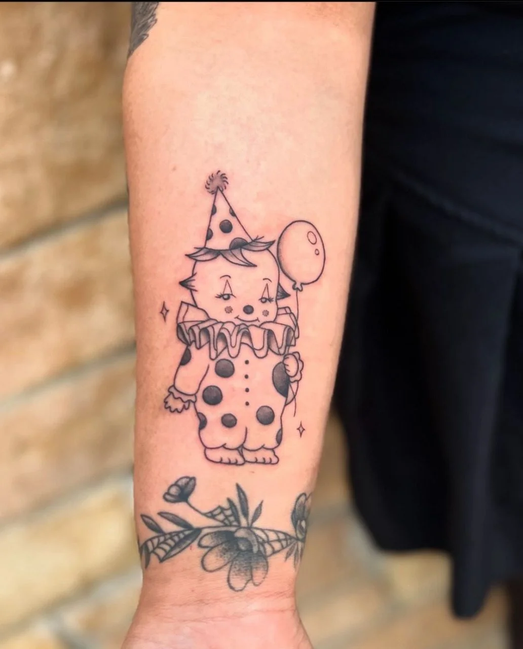 A tattoo of a cute clown dog with a polka-dot costume, ruffled collar, and party hat, holding a balloon, with a floral design below the character.
