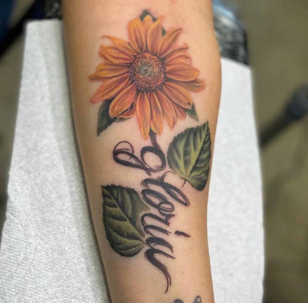 Colorful tattoo of a sunflower with green leaves and black cursive script that reads 'Love' on a person's arm.