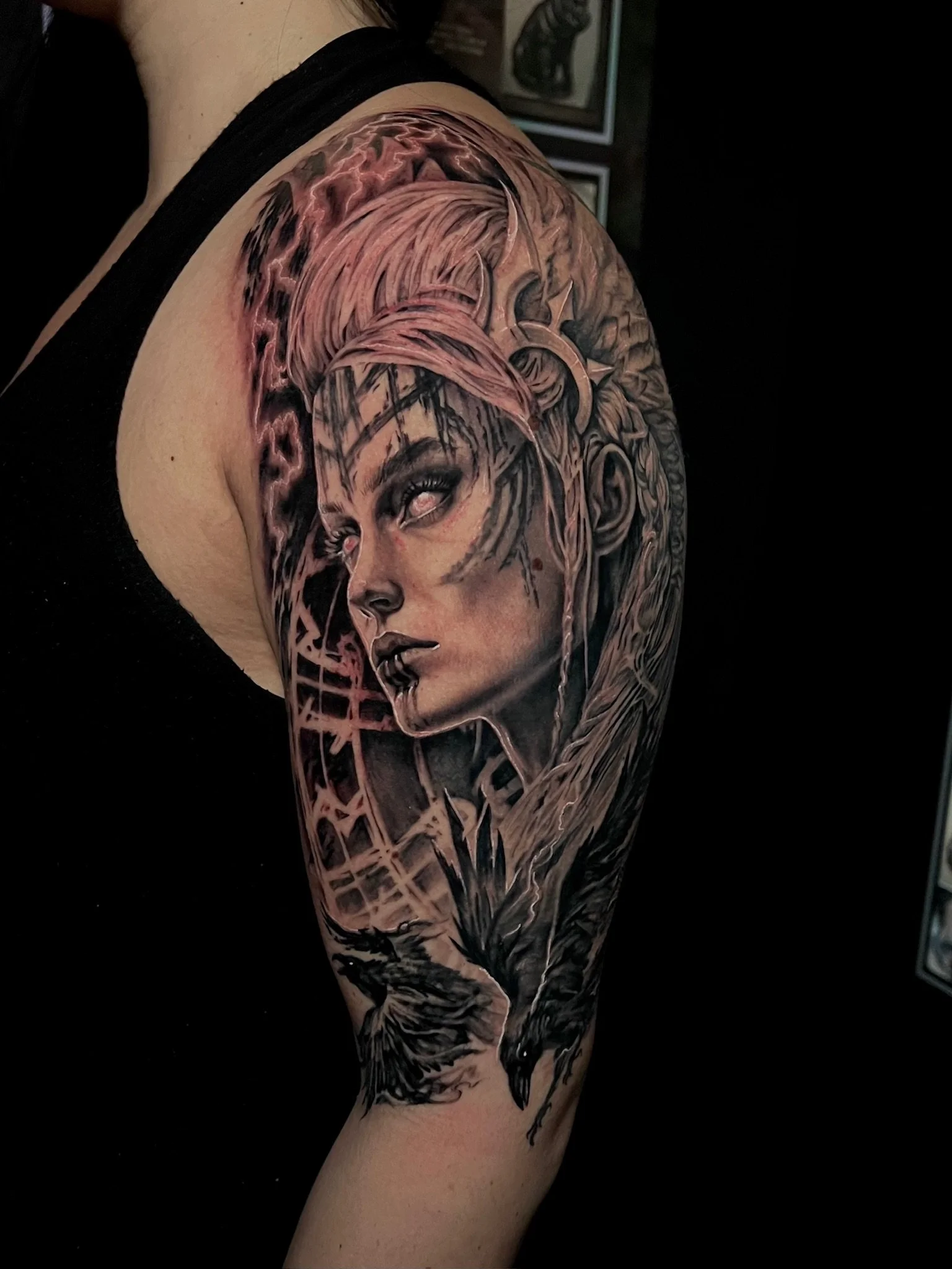 Black and gray tattoo of a woman's face with an elaborate hairstyle, along with two crows, on a person's upper arm.