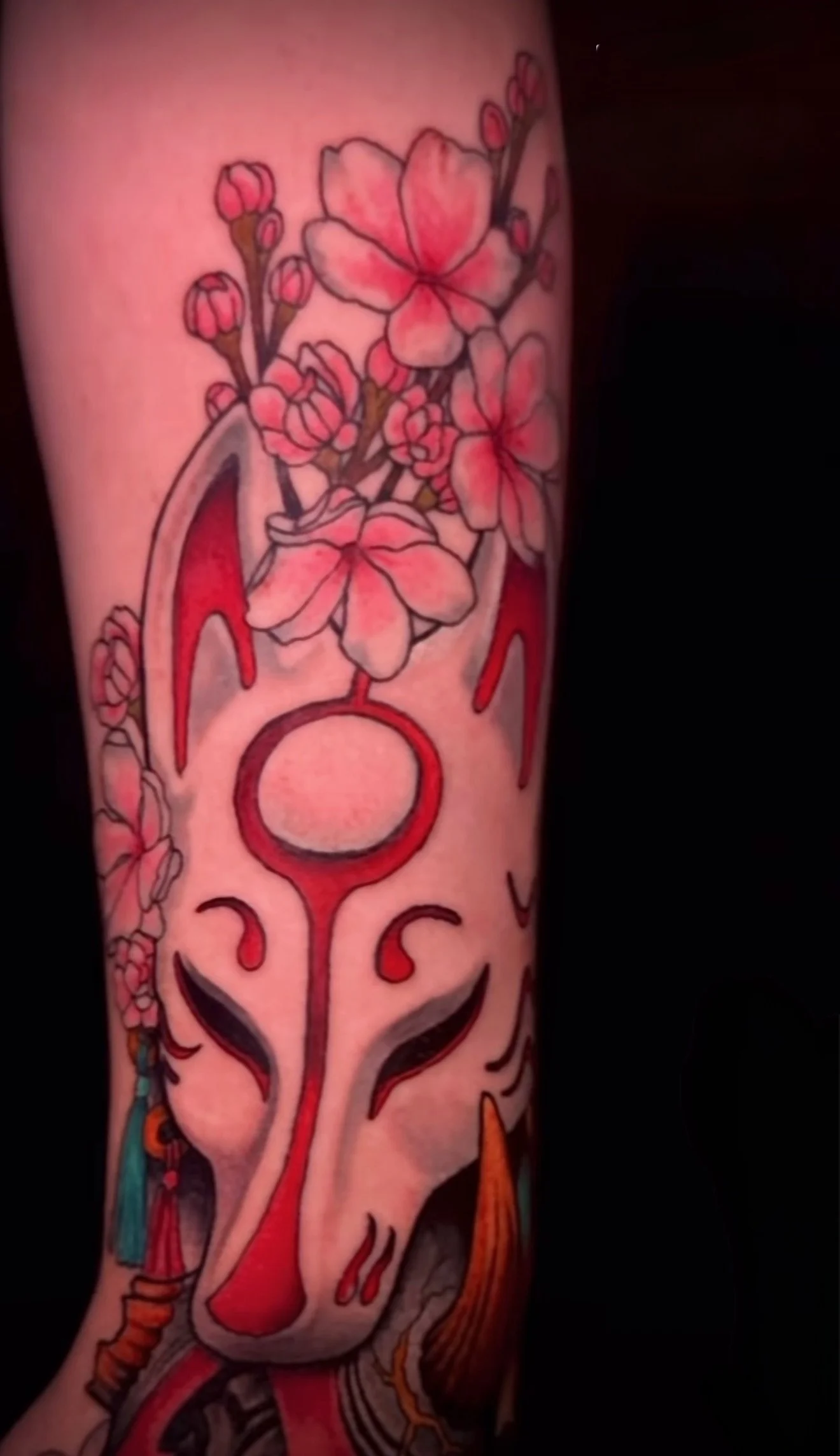 A tattoo of cherry blossoms with pink petals and green leaves, alongside stylized abstract patterns and shapes in red, black, and orange on the forearm.