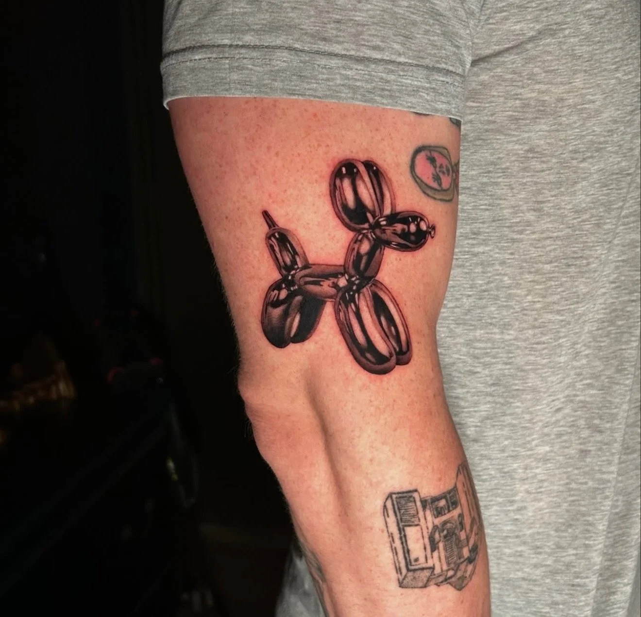 Tattoo of a balloon dog on person's upper arm, with additional tattoos of a small circle with a line and a rectangular object with drawings.
