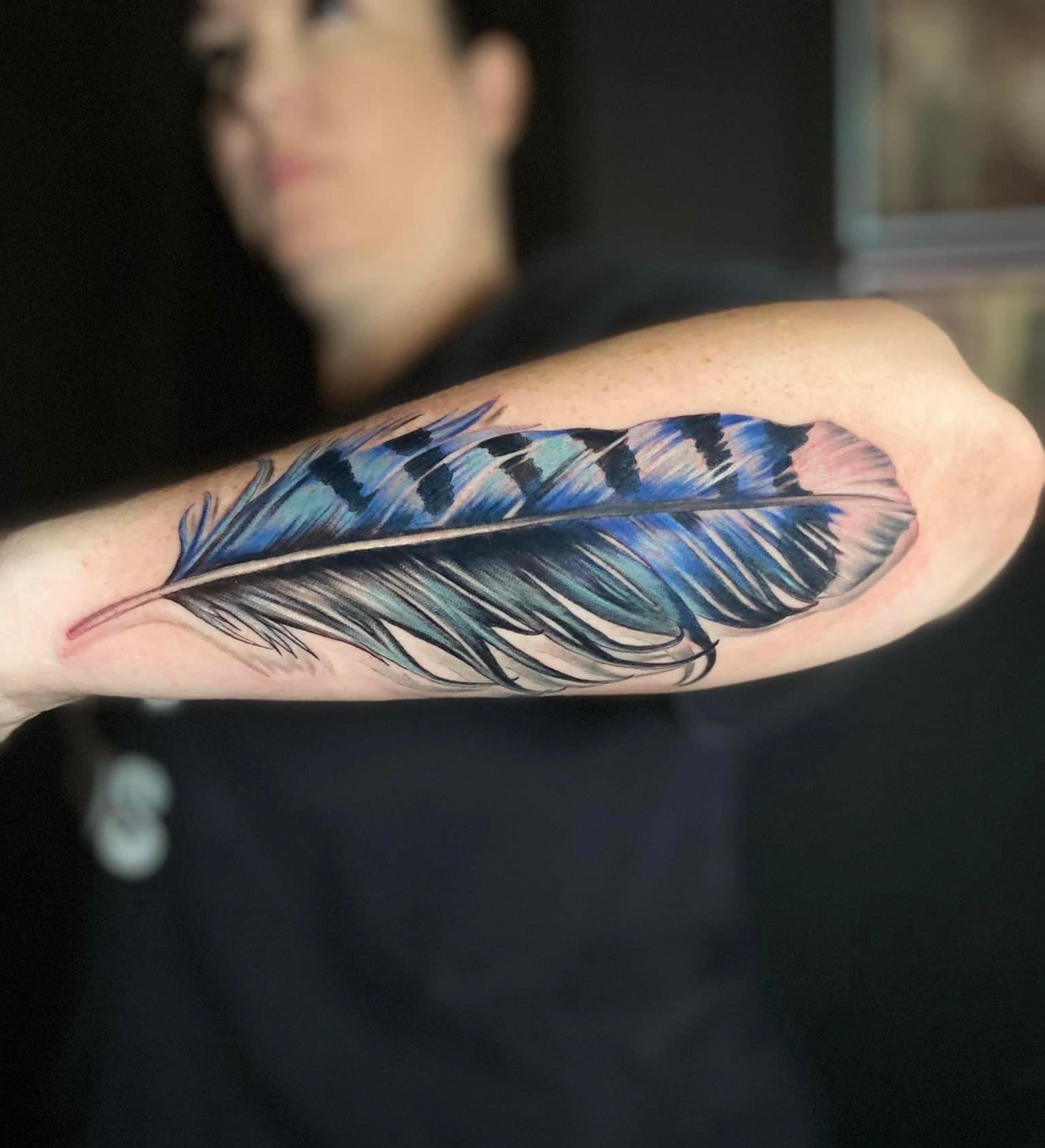 Colorful tattoo of a feather on a person's upper arm, featuring shades of blue, black, and hints of pink.