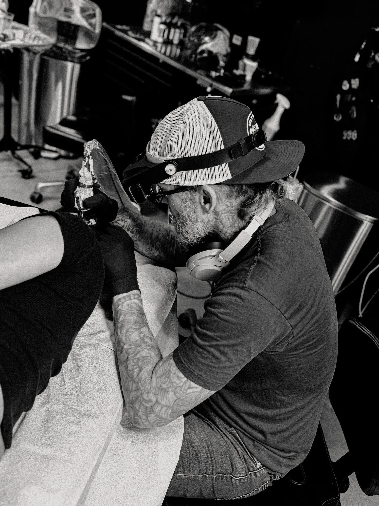 Tattooed man with a beard, wearing a hat, tattooing a person's arm in a tattoo studio.