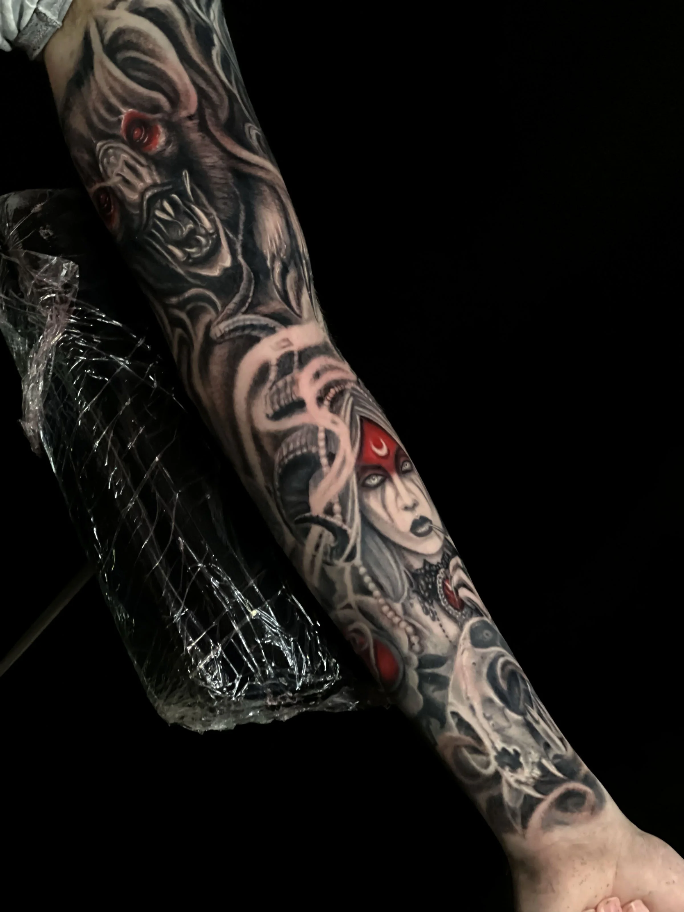Close-up of a person's arm with detailed, colorful tattoos featuring various designs, including a woman's face, a skull, and other intricate elements, against a black background.