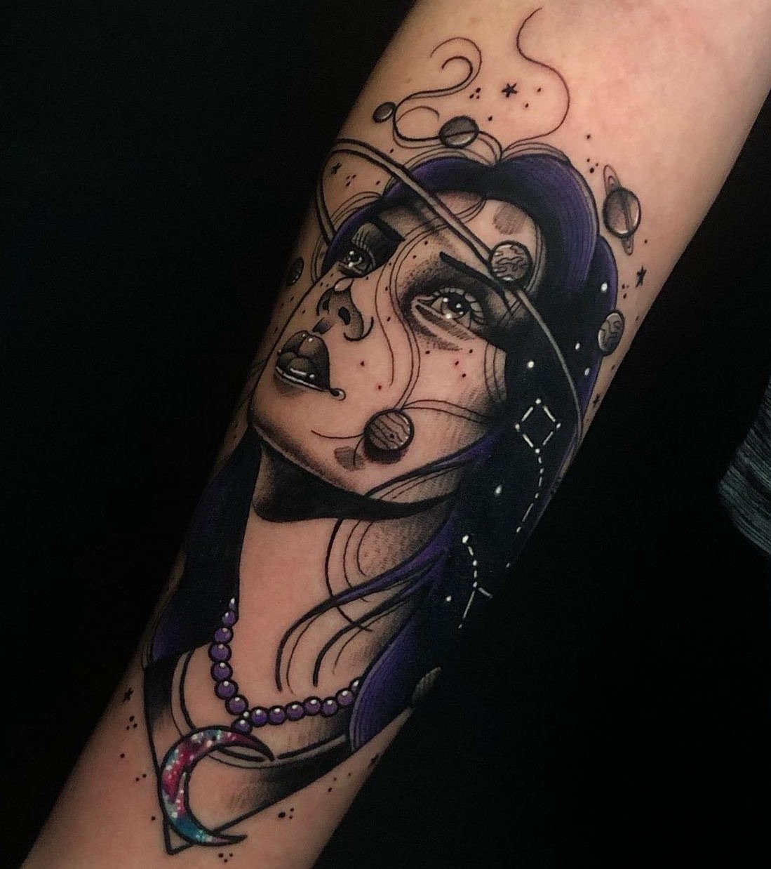 Tattoo of a woman's face with cosmic elements like planets, stars, and swirling lines, featuring purple hair, a beaded necklace, and a crescent moon pendant.