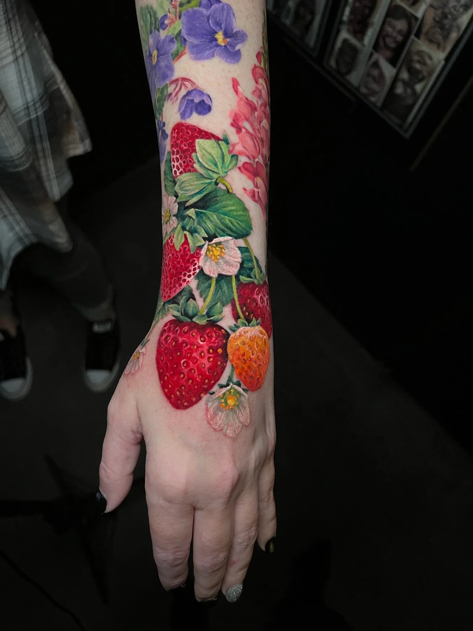 Colorful tattoo of strawberries, strawberries flowers, and purple violets on a person's forearm and hand.