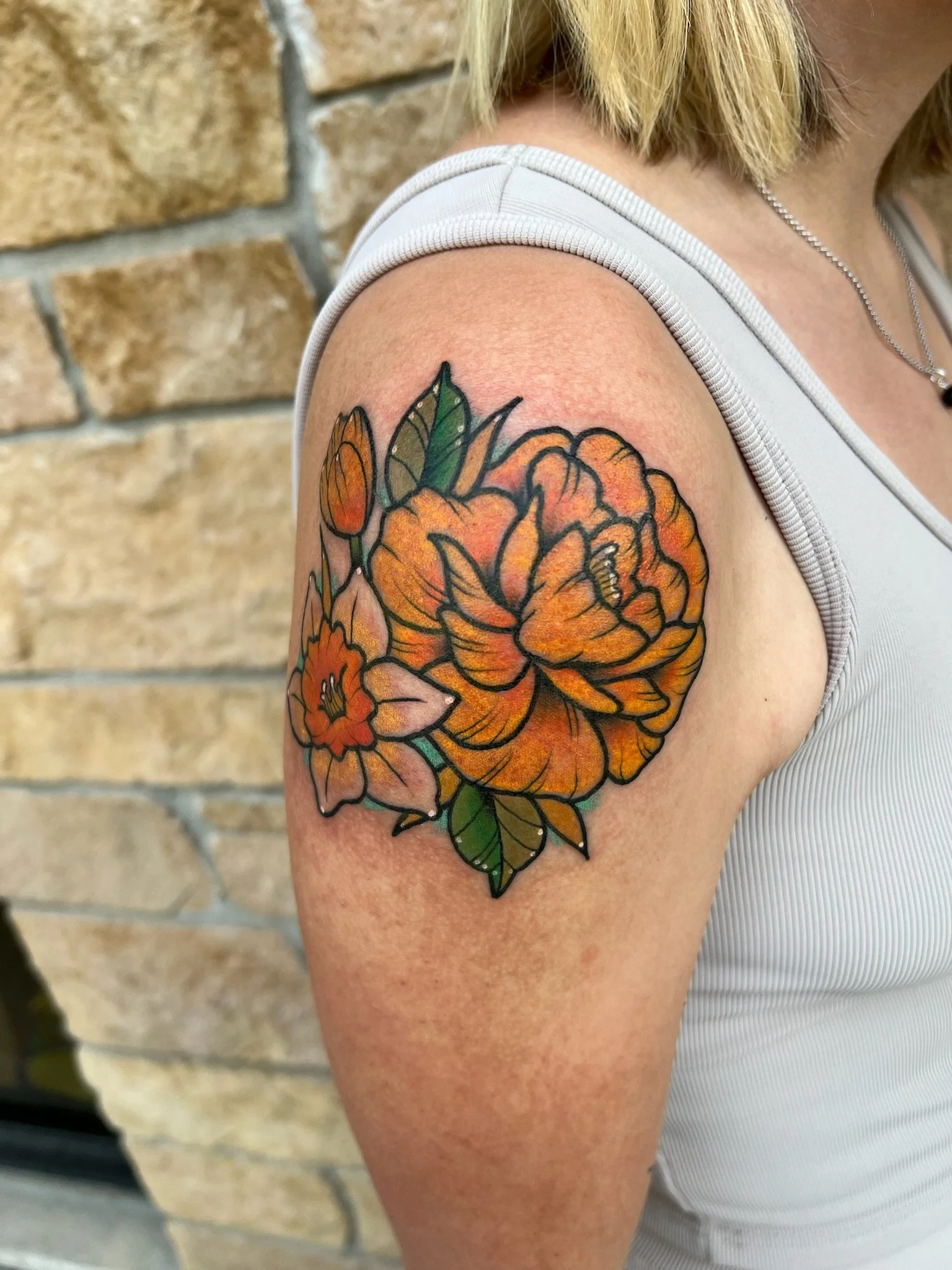 Colorful floral tattoo on person's upper arm featuring large orange and pink flowers, with green leaves, outlined in black.