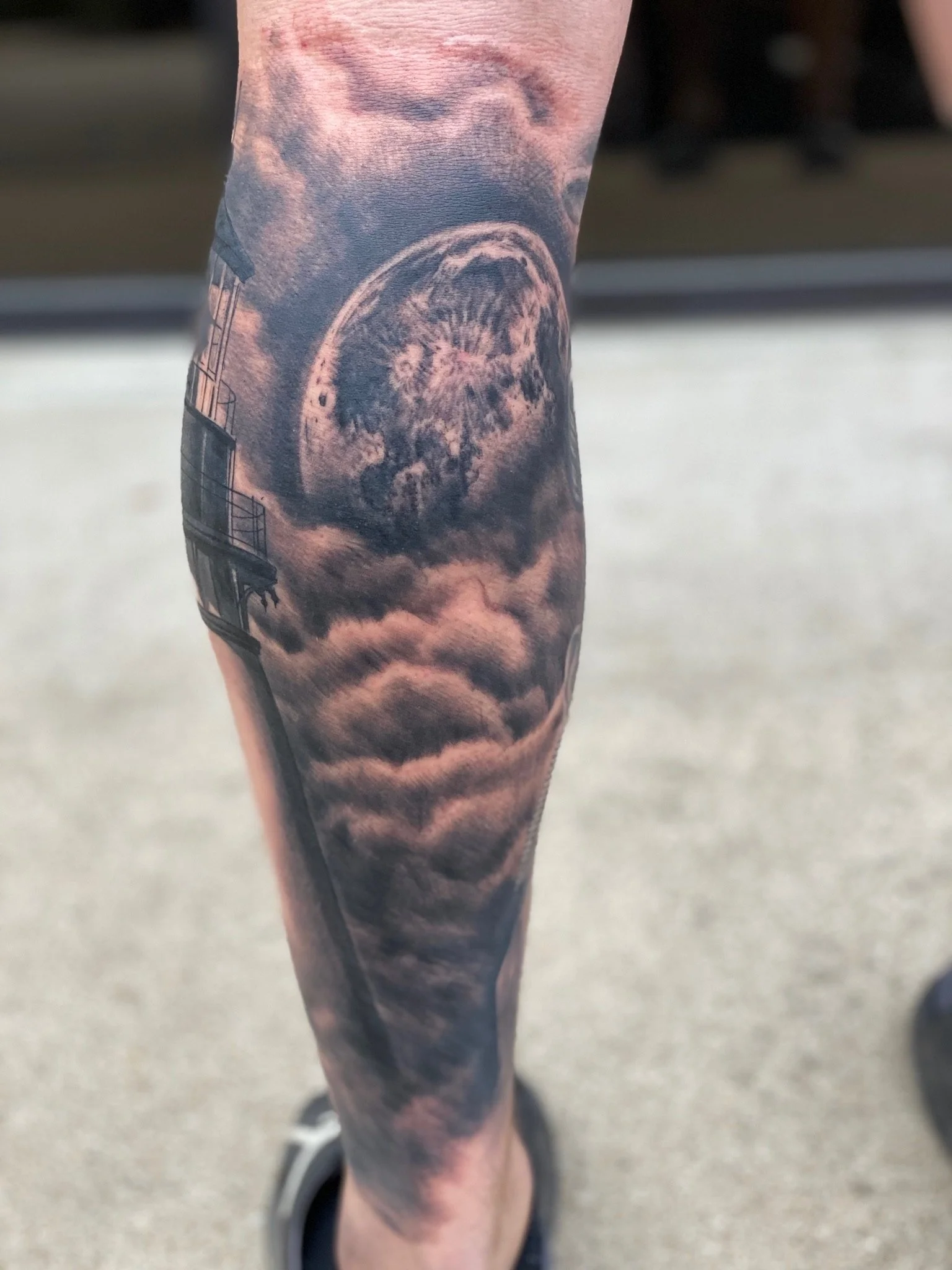 Tattoo of a moon surrounded by clouds on a person's lower leg.