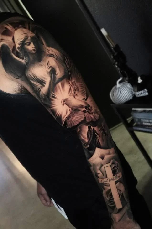 Close-up of a person's arm with a detailed black and gray tattoo sleeve featuring a woman with long hair, a dove, roses, and a cross with the words 'God Will'.