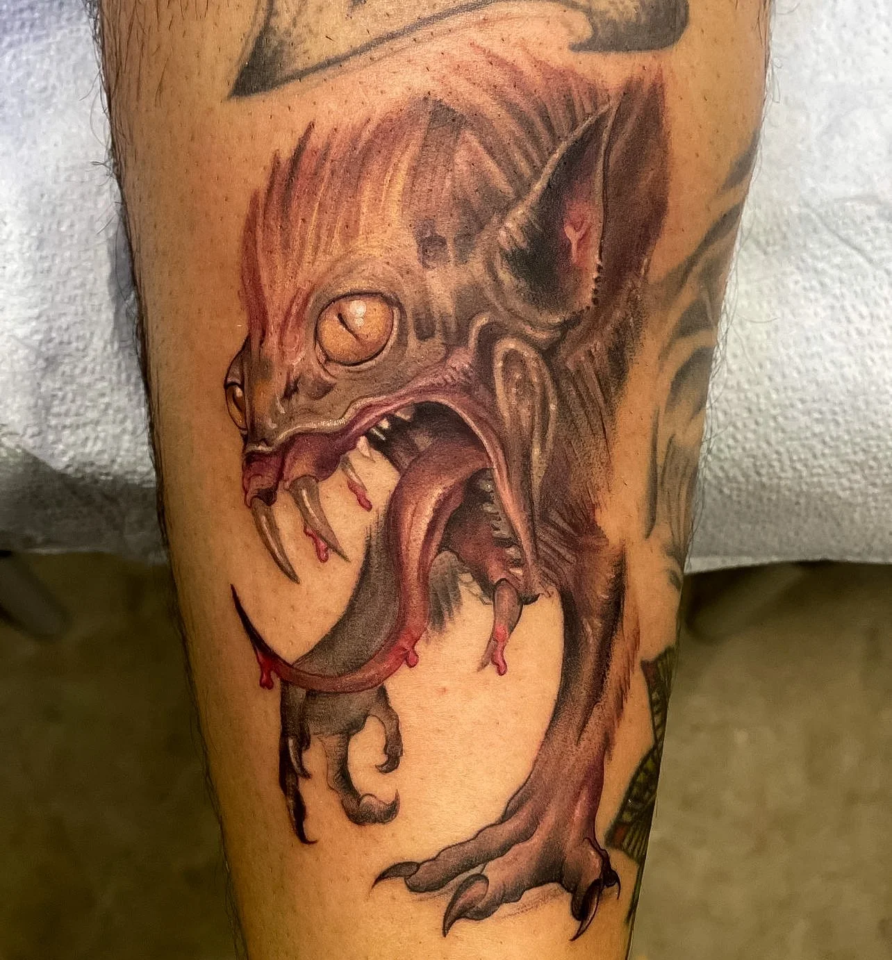 Tattoo of a menacing, exaggerated bat with open mouth showing sharp teeth and blood dripping, rendered in dark, realistic style on a person's arm.