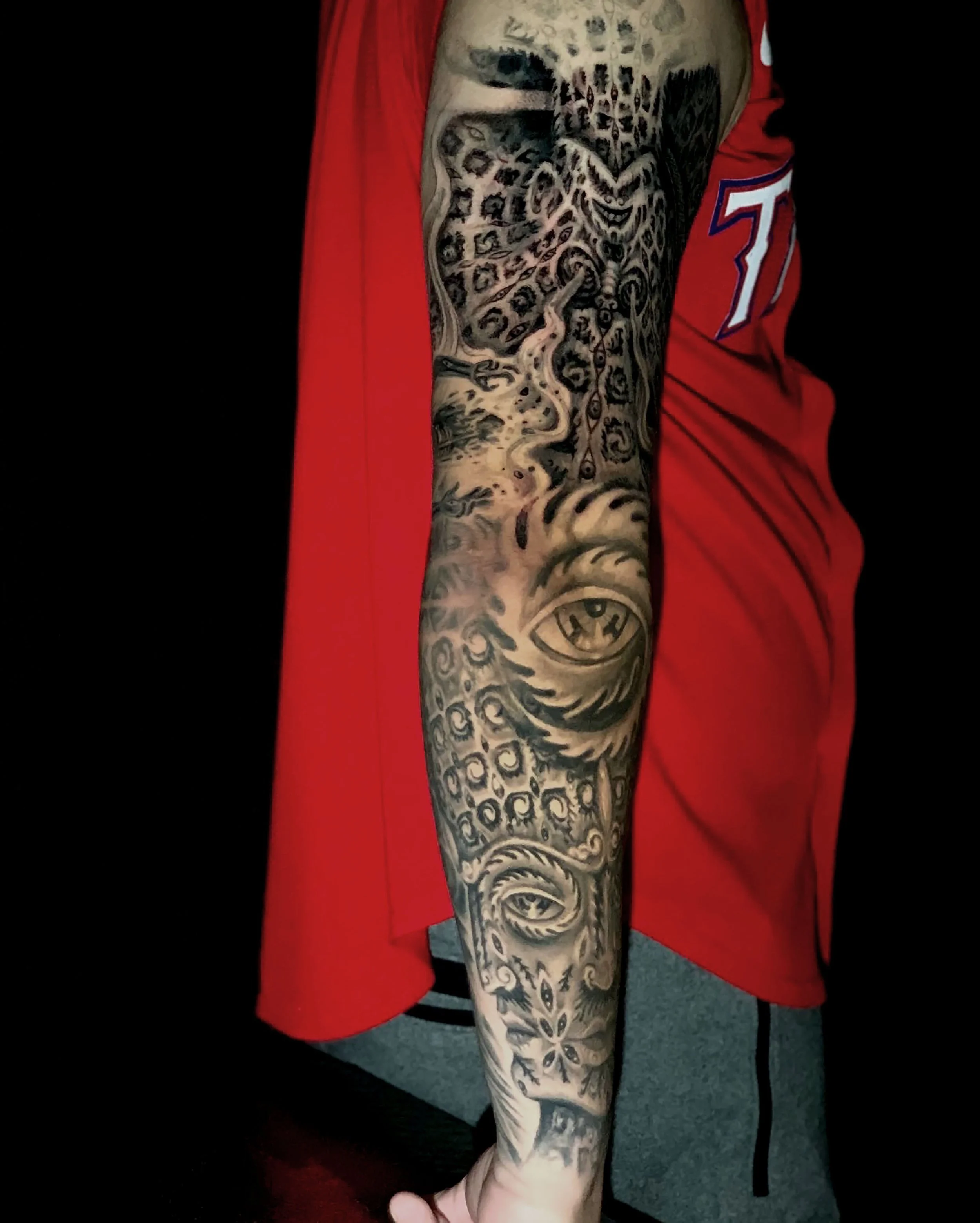 Close-up of a person's arm covered in detailed black and gray tattoo sleeve with various intricate designs, including a large eye near the wrist, a face, and floral patterns, against a red shirt and gray shorts.