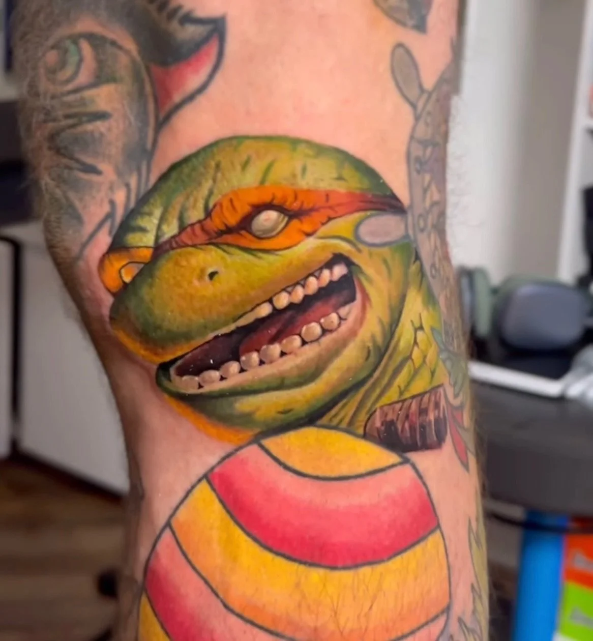 Colorful tattoo of a cartoonish, smiling dinosaur or reptile on a person's arm, with detailed scales and markings.