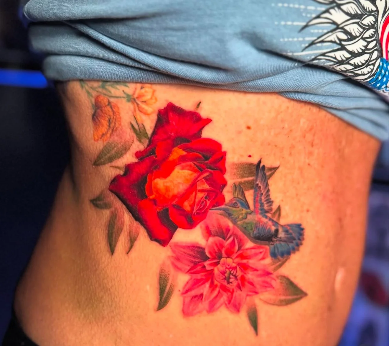 A colorful tattoo of a red rose, a pink flower, and a hummingbird, with green leaves, on a person's skin.