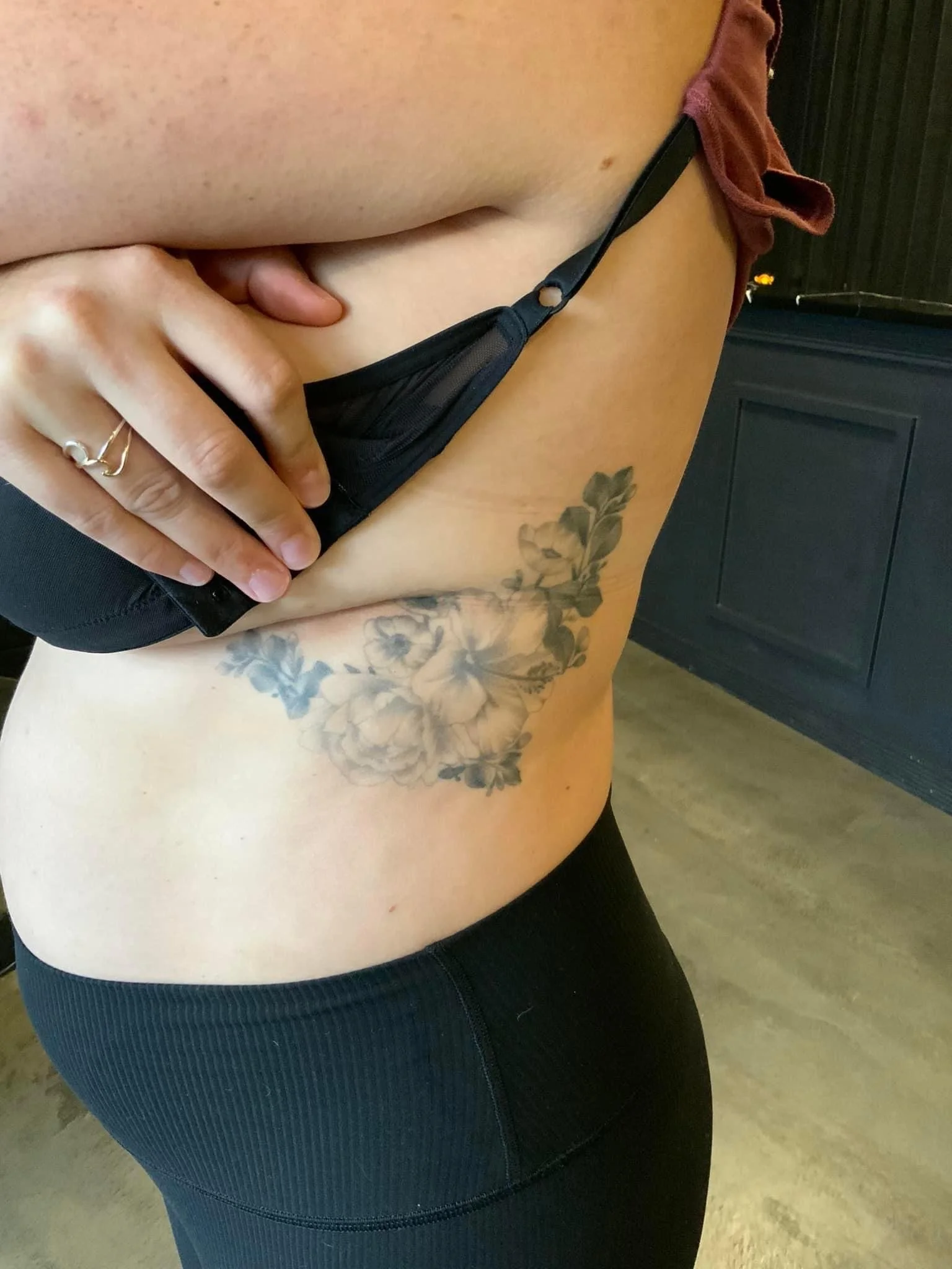A woman with a floral tattoo on her side is lifting her shirt to reveal it.